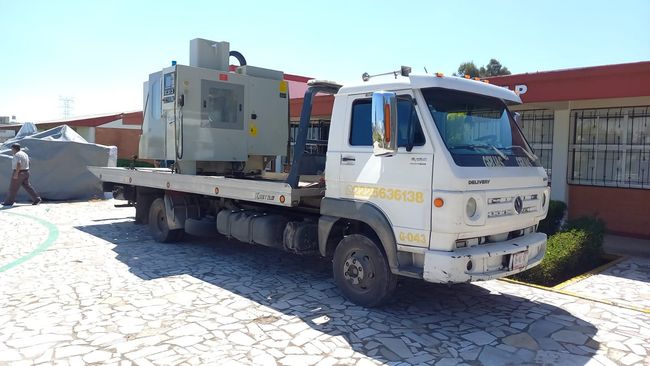 Camión de plataforma que transporta maquinaria industrial de gran tamaño estacionado frente a un edificio.