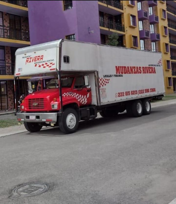 Camión de mudanzas rojo y blanco de Mudanzas Rivera en una calle, estacionado frente a un edificio de apartamentos.