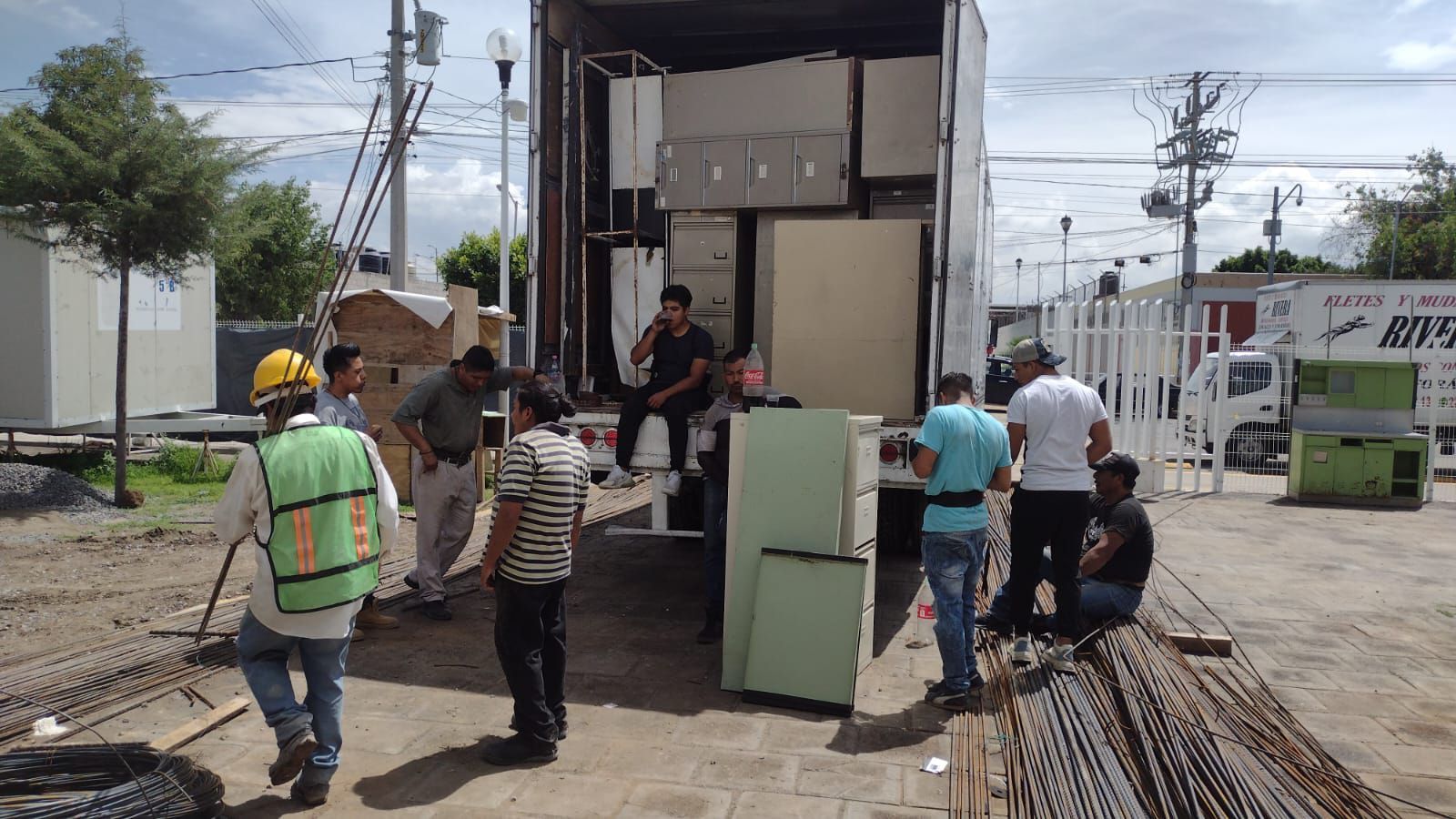 Trabajadores descargando muebles de un camión al aire libre. Algunos llevan chalecos de seguridad y cascos.
