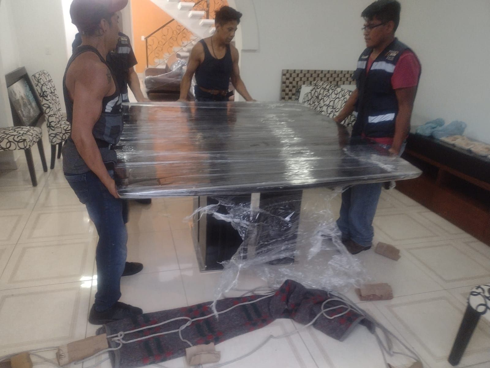 Cuatro hombres levantan una gran mesa envuelta en negro dentro de una habitación.