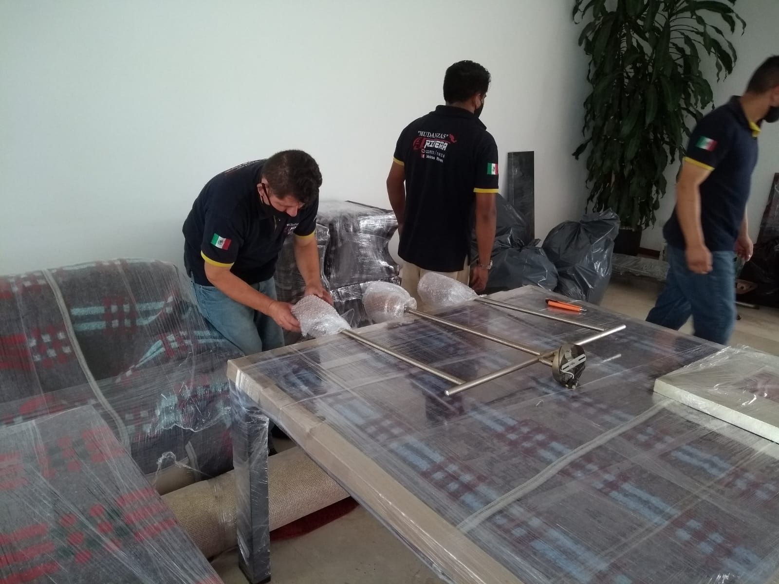 Mudanzas envolviendo muebles, una mesa y otros artículos dentro de una casa. Tres hombres llevan mascarillas y camisas azules a juego.