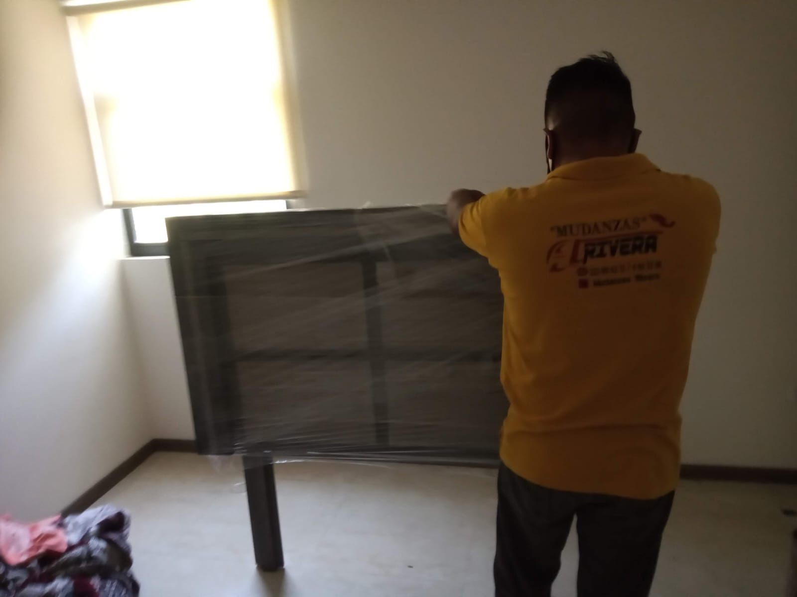 Hombre con camisa amarilla envolviendo un objeto negro con film plástico en una habitación vacía cerca de una ventana.