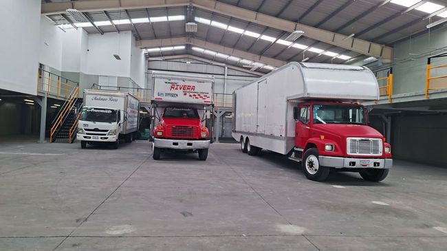 Tres camiones estacionados dentro de un almacén: blanco, rojo y rojo, con remolques en la parte superior de color blanco.