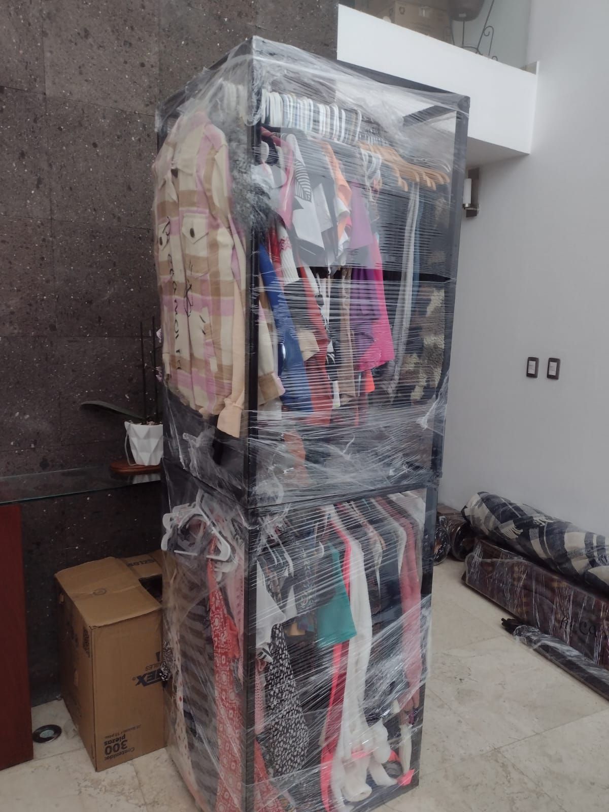 Perchero, apilado y envuelto en plástico, que contiene diversas prendas en un espacio interior.