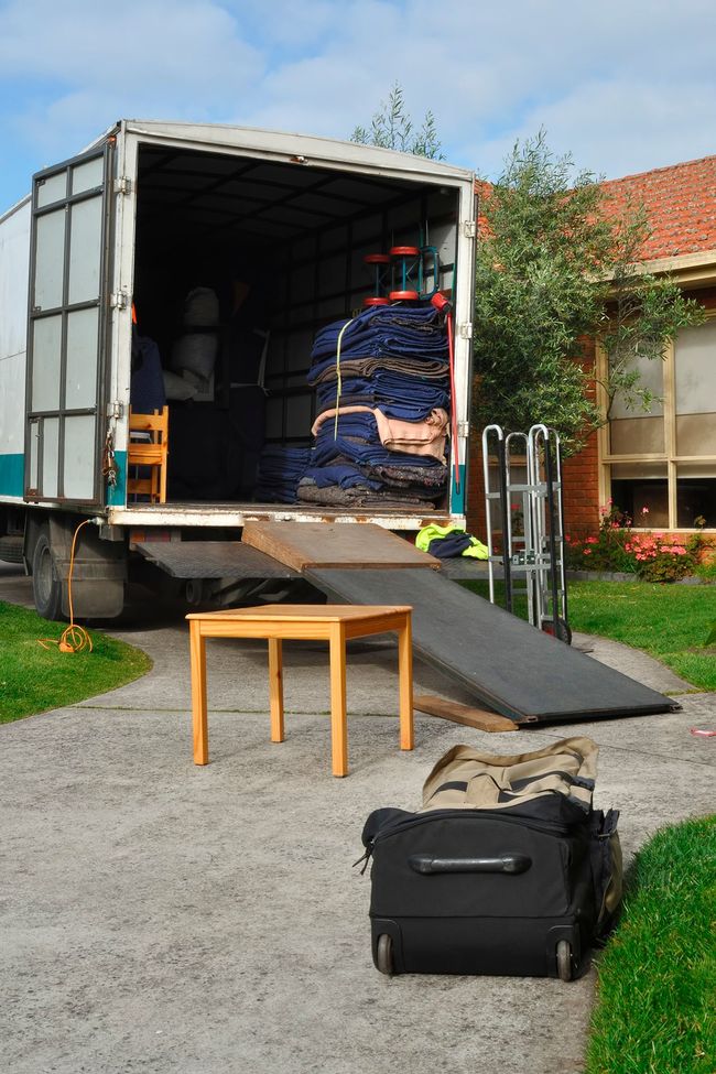 Camión de mudanzas estacionado en la entrada, rampa de carga hacia abajo; muebles y equipaje estacionados cerca.