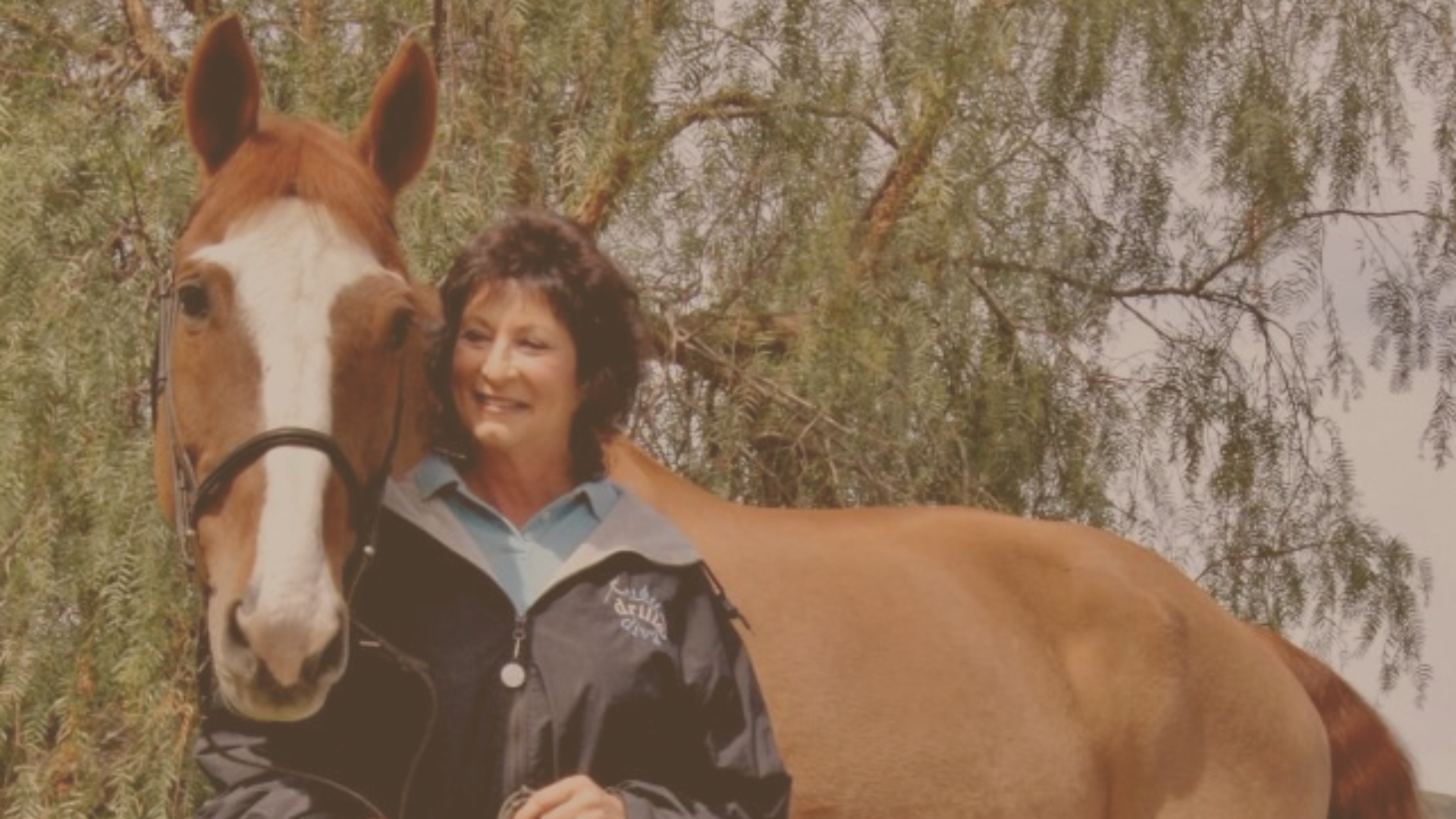 smiling brunette woman wearing dark jacket standing close to a brown horse with white stripe on nose