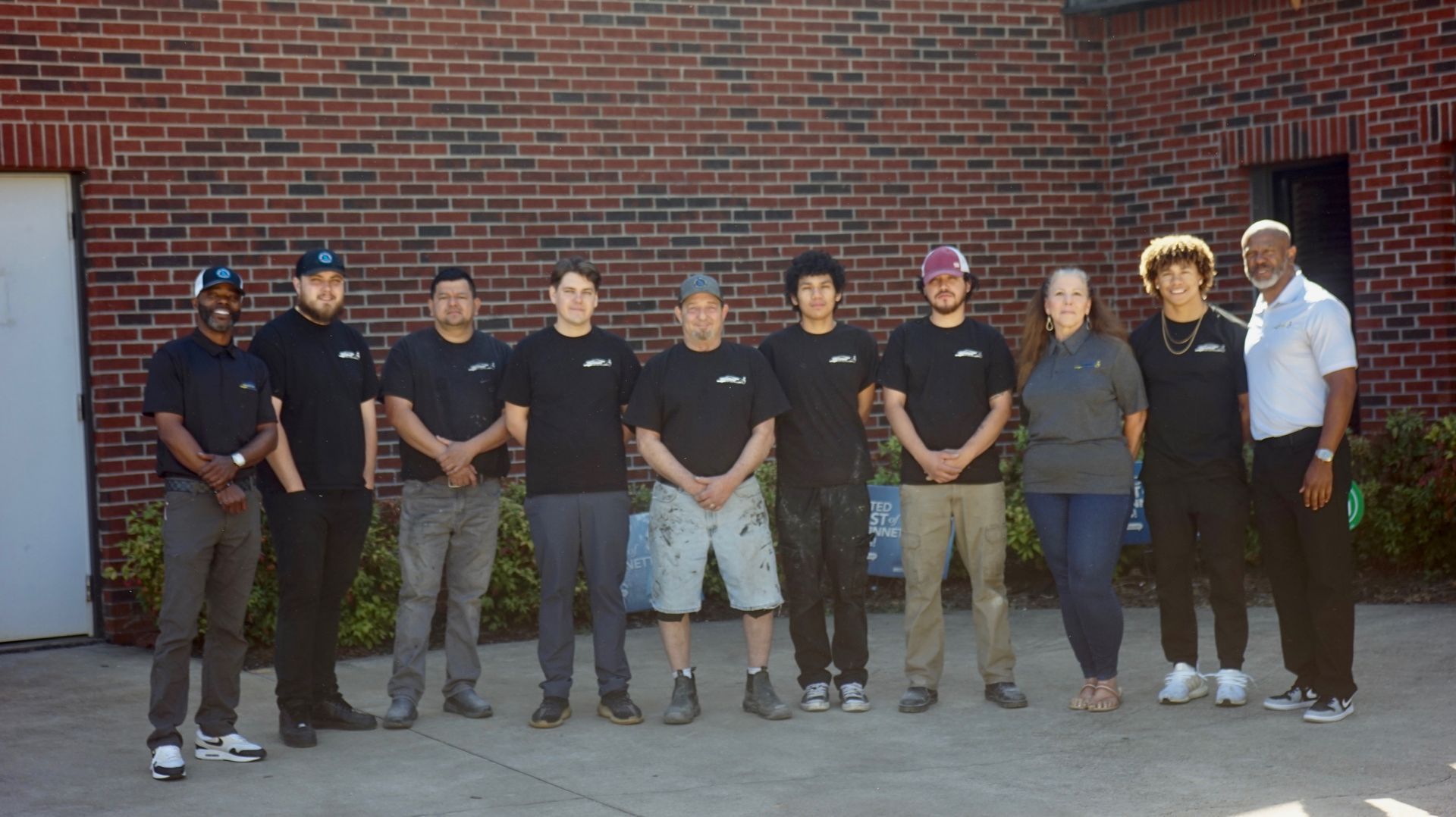 Eight people stand in a row outside a brick building, wearing mostly black shirts and jeans.