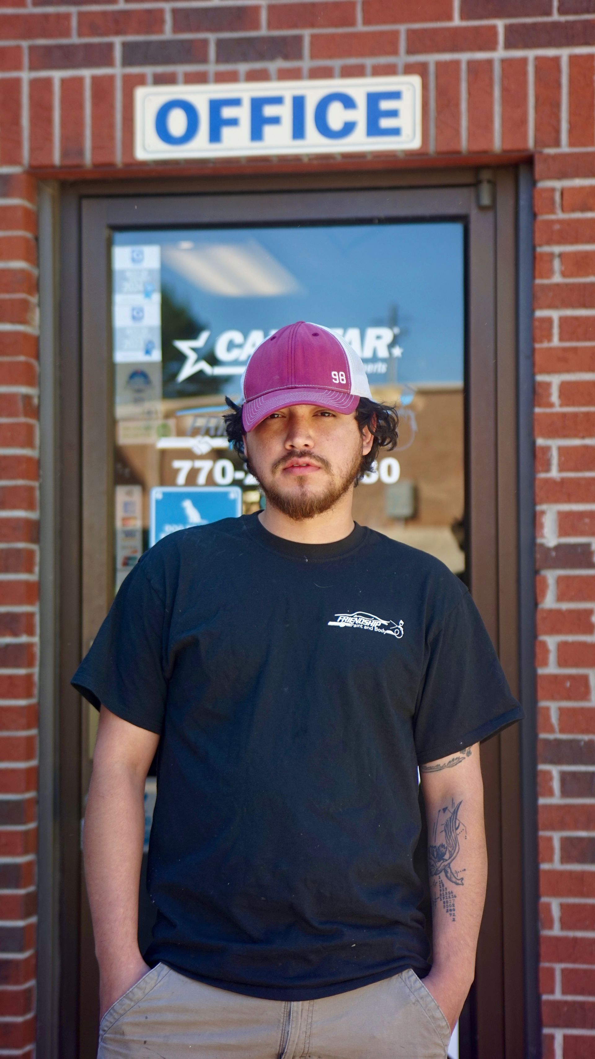 Person standing outside an office doorway, wearing a black T-shirt and pink cap, in front of a brick building.