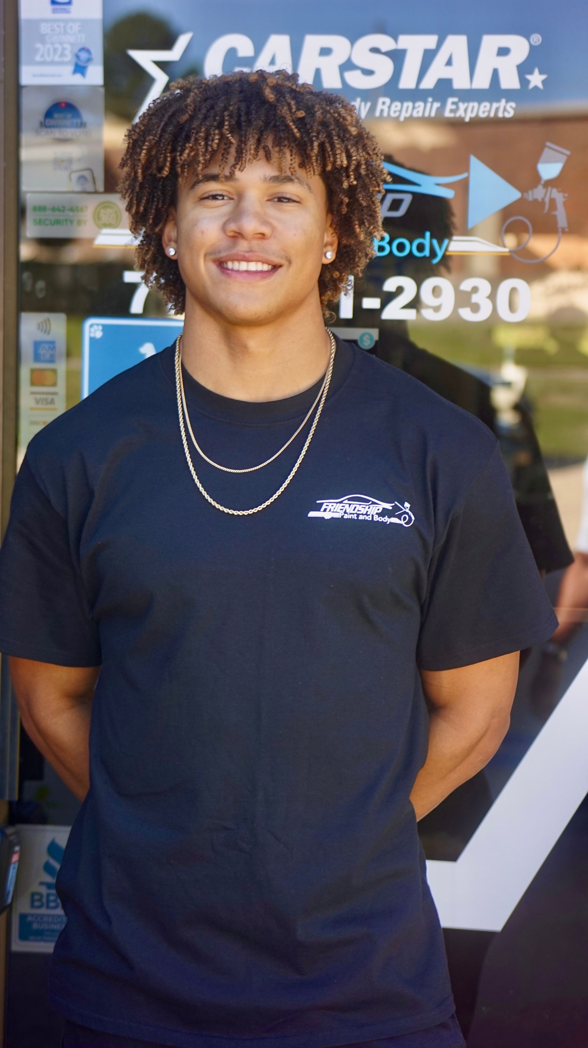 Person smiling in front of a CARSTAR auto repair sign, wearing a black T-shirt and gold chains