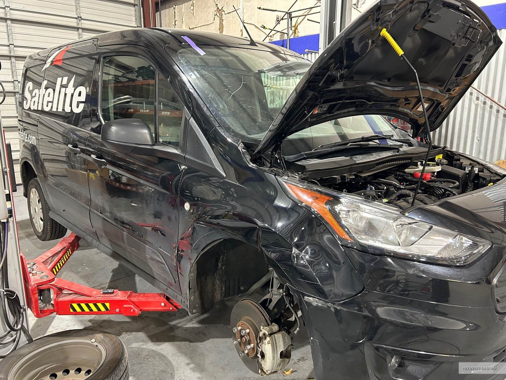 Black minivan with front-end damage and hood open in a garage, wheel removed, sat on red jack stands