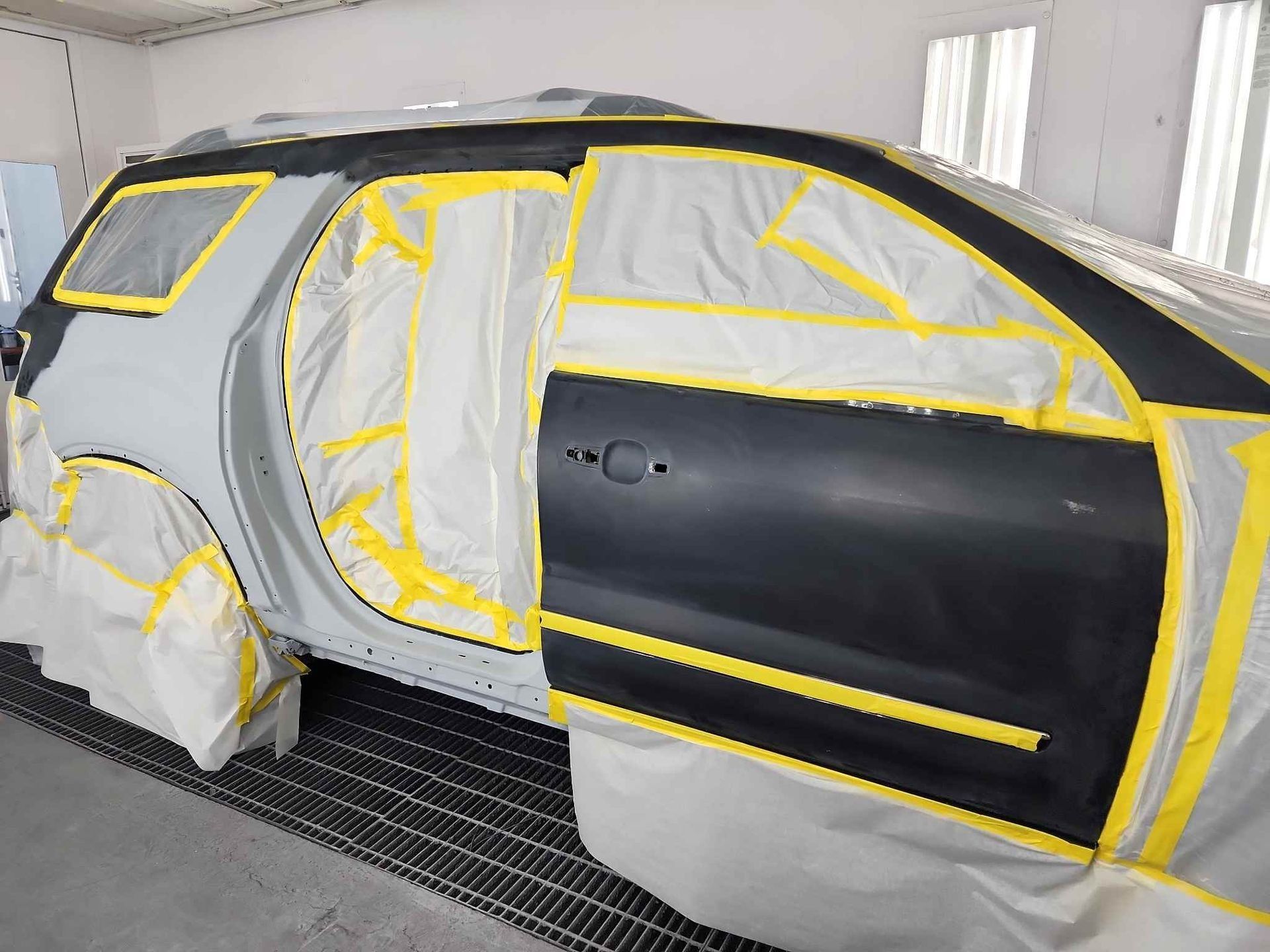 Car body masked with yellow tape and white paper in a paint booth, black door panel visible