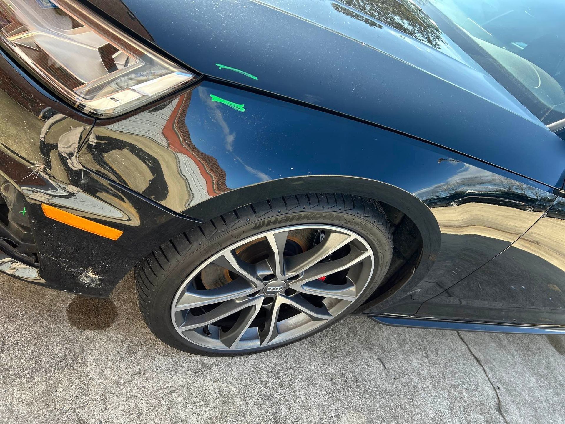 Close-up of a black car’s front fender and wheel with green and orange paint scratches.