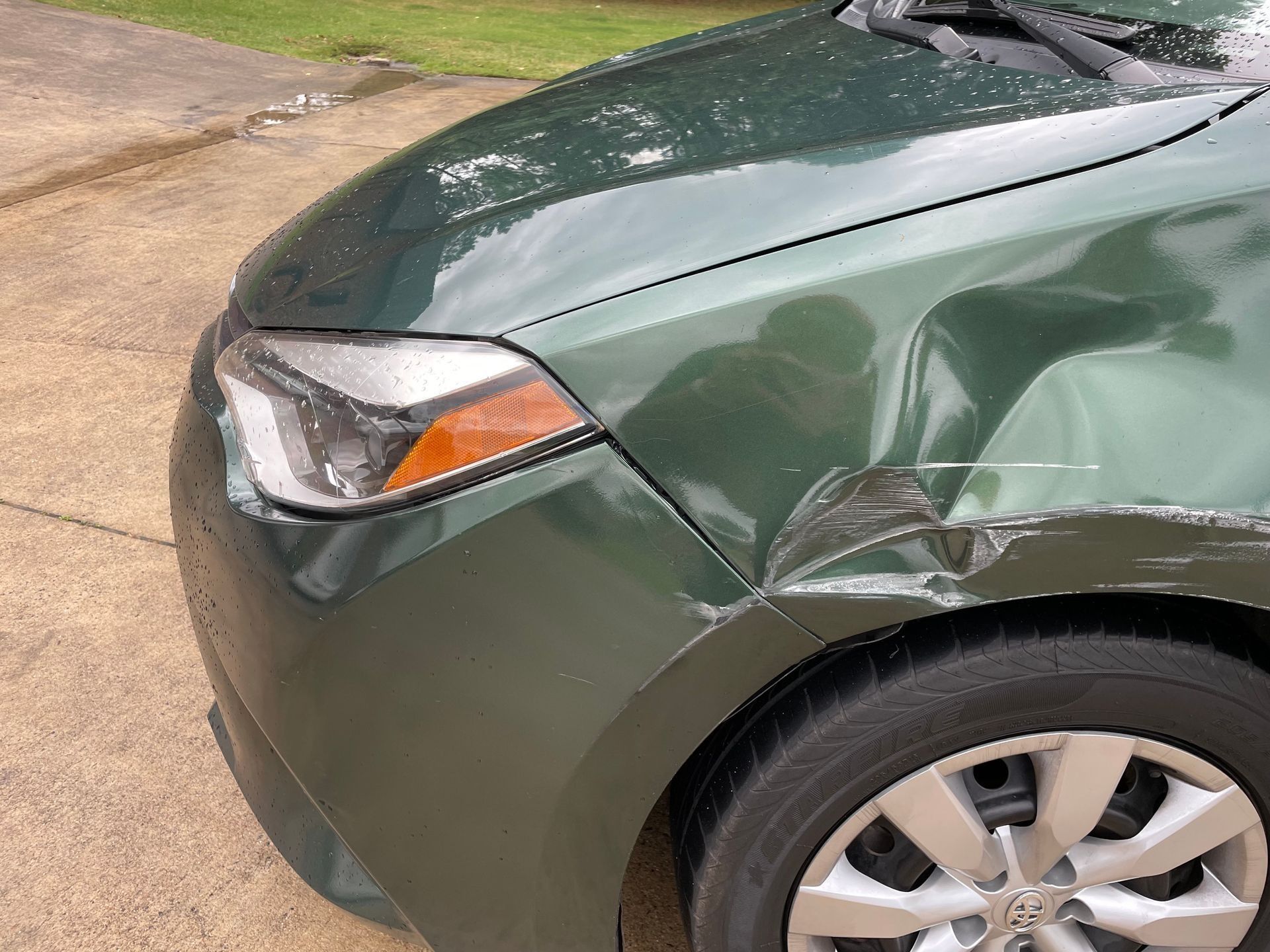 Front left side of a green car with a dented fender and scratched hood.
