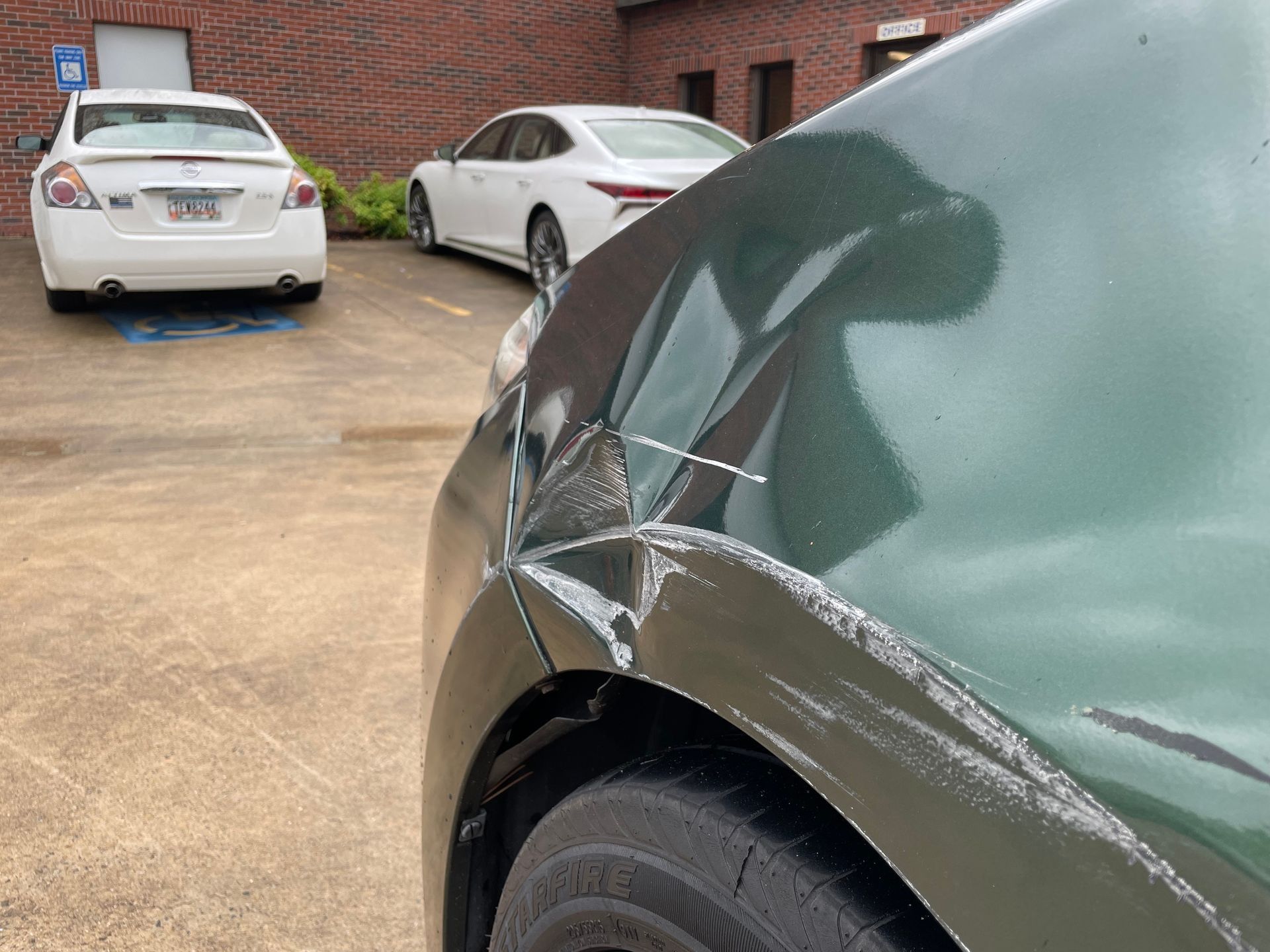 Damaged front fender on a green car in a parking lot, with two white cars nearby.