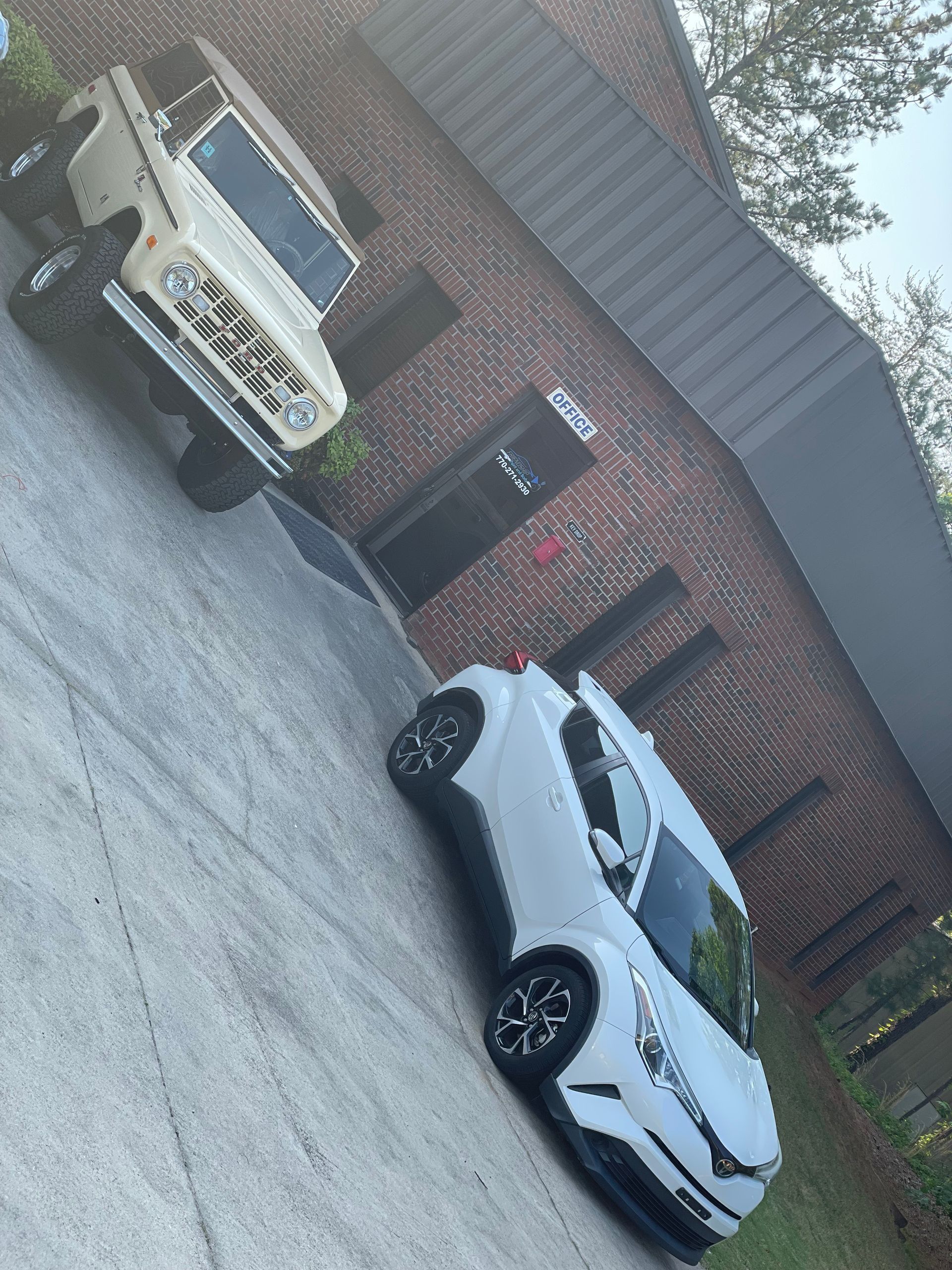 Two parked cars, one white and one beige, outside a brick house on a driveway.