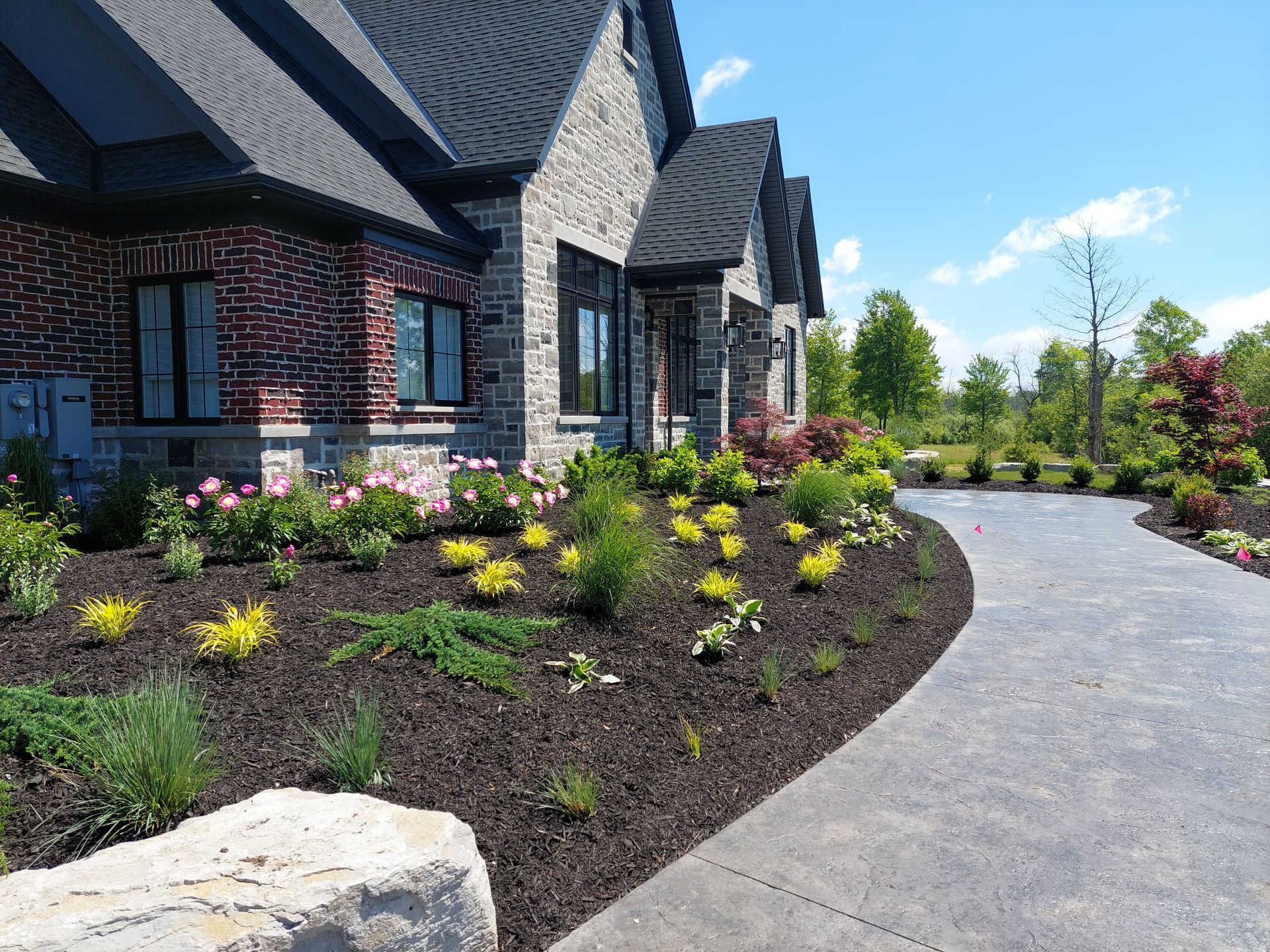 A stone walkway curves through a landscaped garden bed with dark mulch and various plants in front of a modern brick home.
