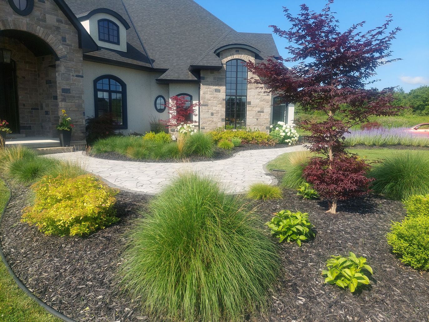 A stone house with dark trim behind a landscaped yard featuring black mulch, ornamental grasses, and a purple-leaf tree.