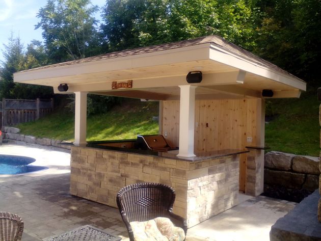 Outdoor bar with a stone base and wooden roof by a swimming pool, featuring integrated speakers and seating.