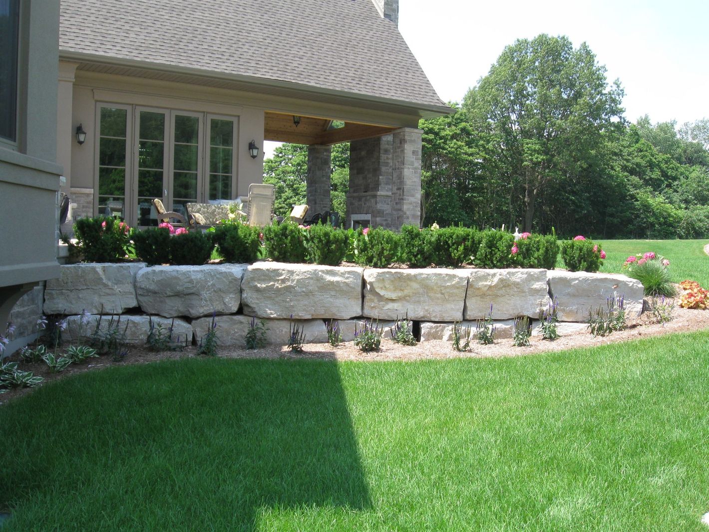 A stone retaining wall sits in a lush green backyard, fronted by a row of small shrubs and a house with a covered patio.