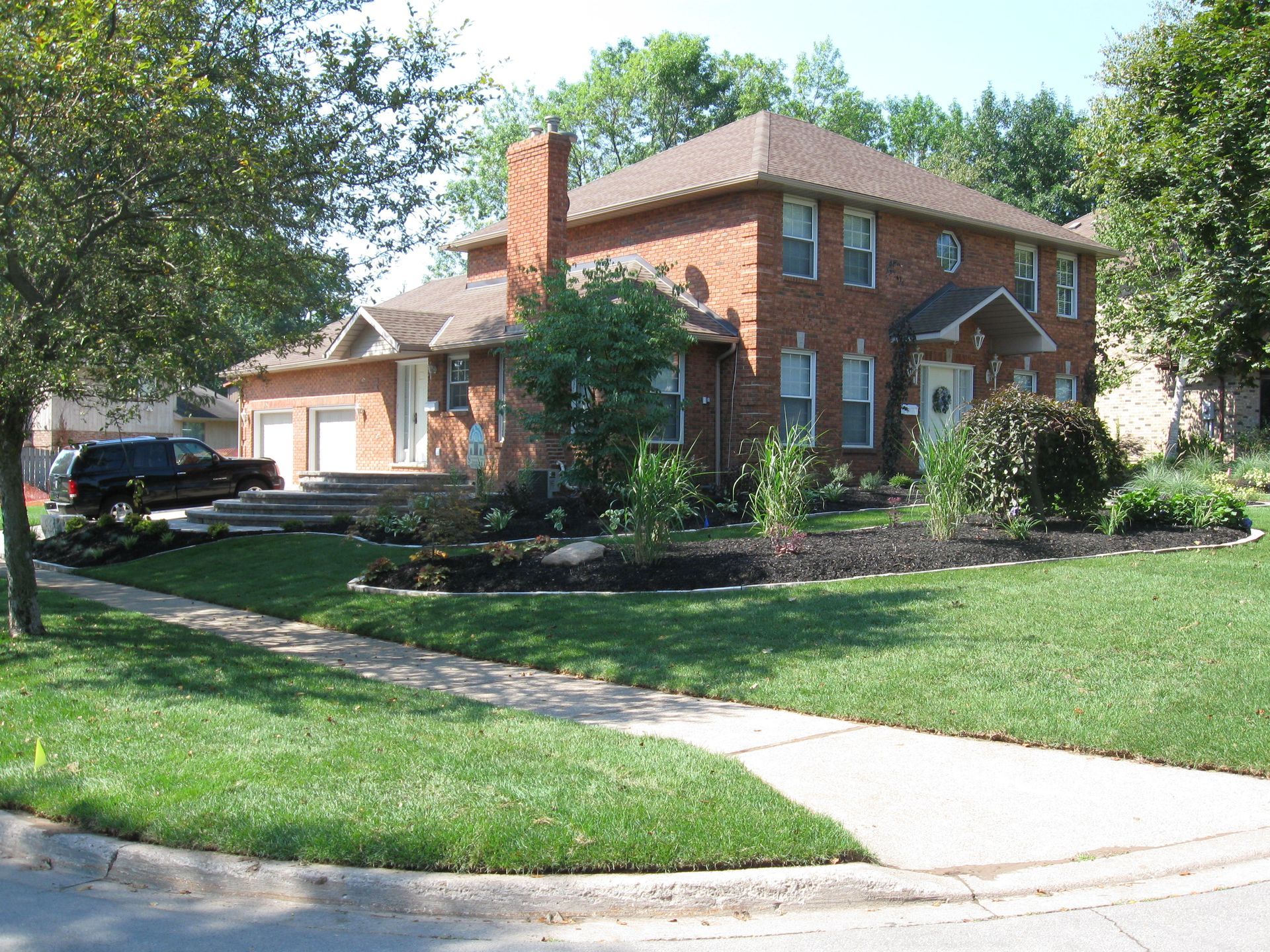 A two-story red brick house with a dark roof and chimney, featuring a landscaped front yard and a concrete driveway.