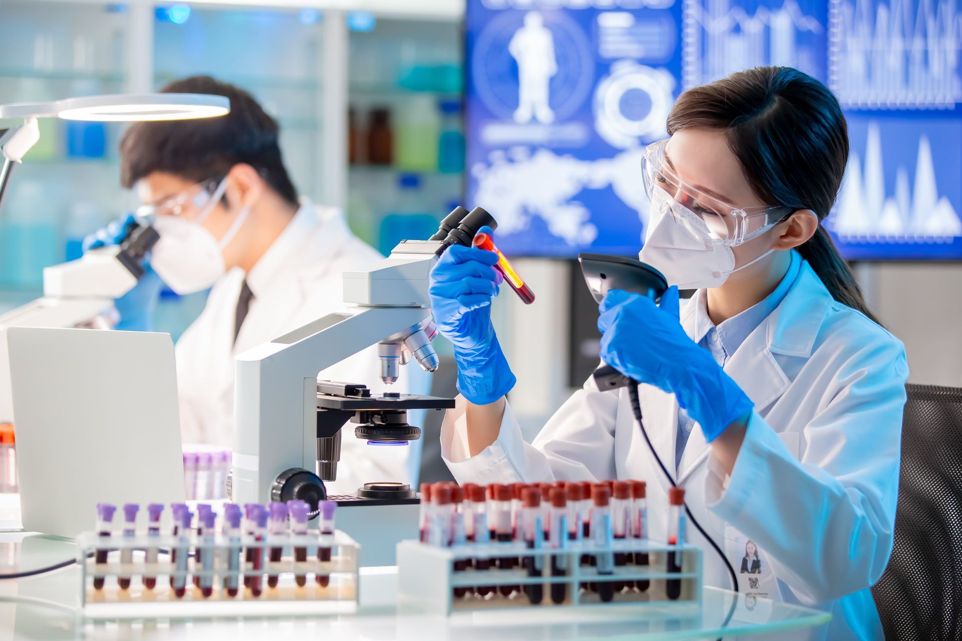 Two lab technicians in masks and gloves pipette samples in a bright medical lab.