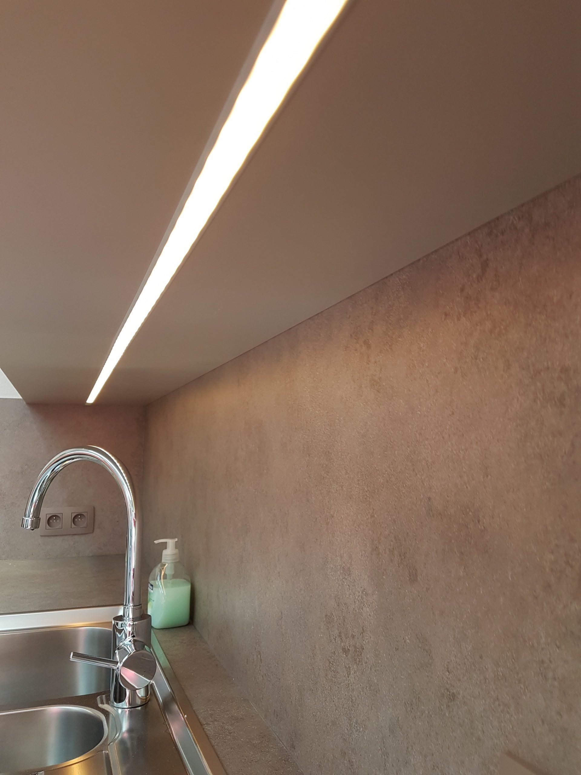 Keuken met een spoelbak, kraan en een ledstrip die het aanrechtblad en de spatwand verlicht. De muren hebben een bruine, getextureerde afwerking.