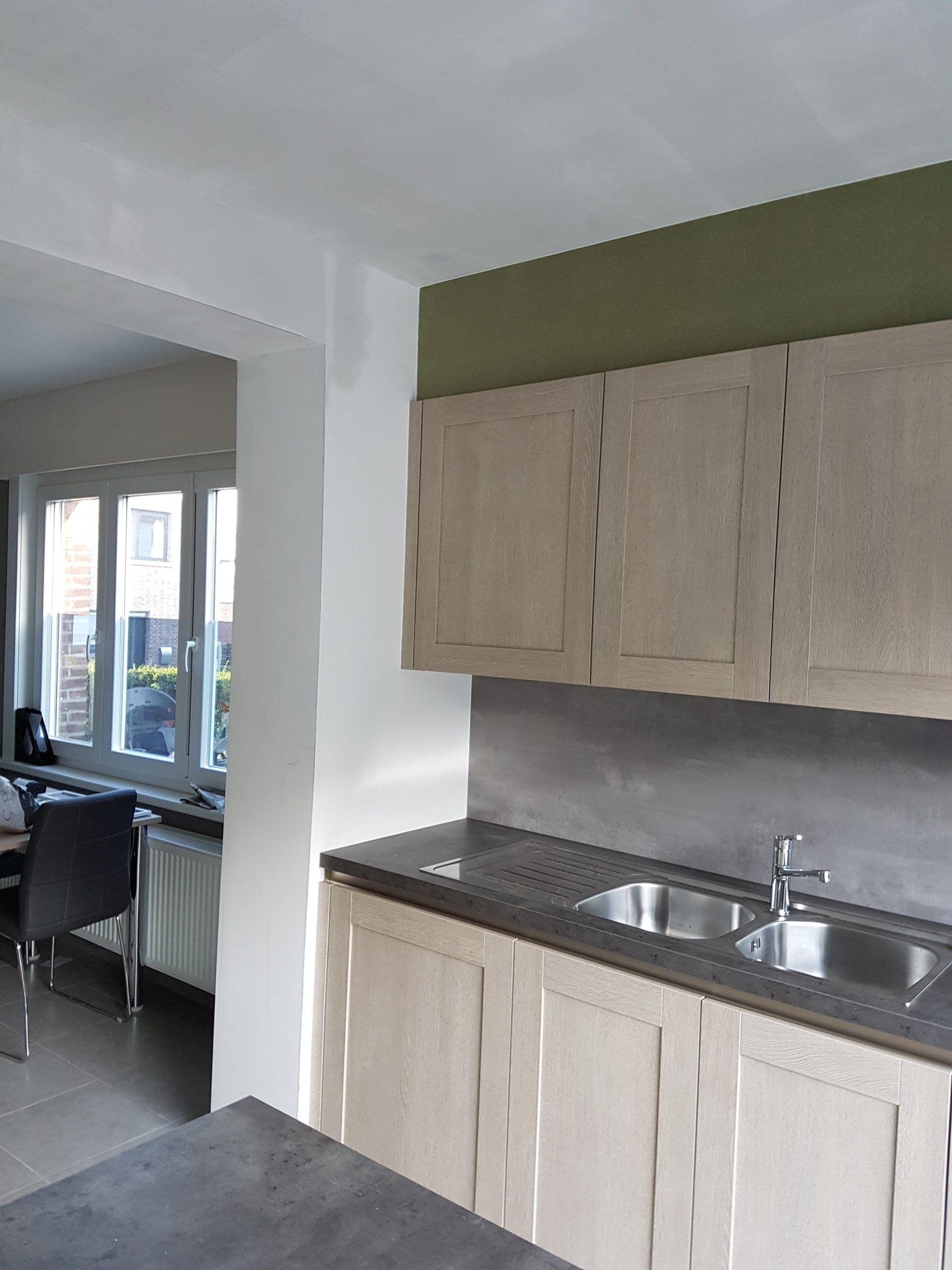 Keuken met lichthouten kasten, een grijs aanrechtblad en een roestvrijstalen spatwand. Een olijfgroene muur zorgt voor een vleugje kleur.