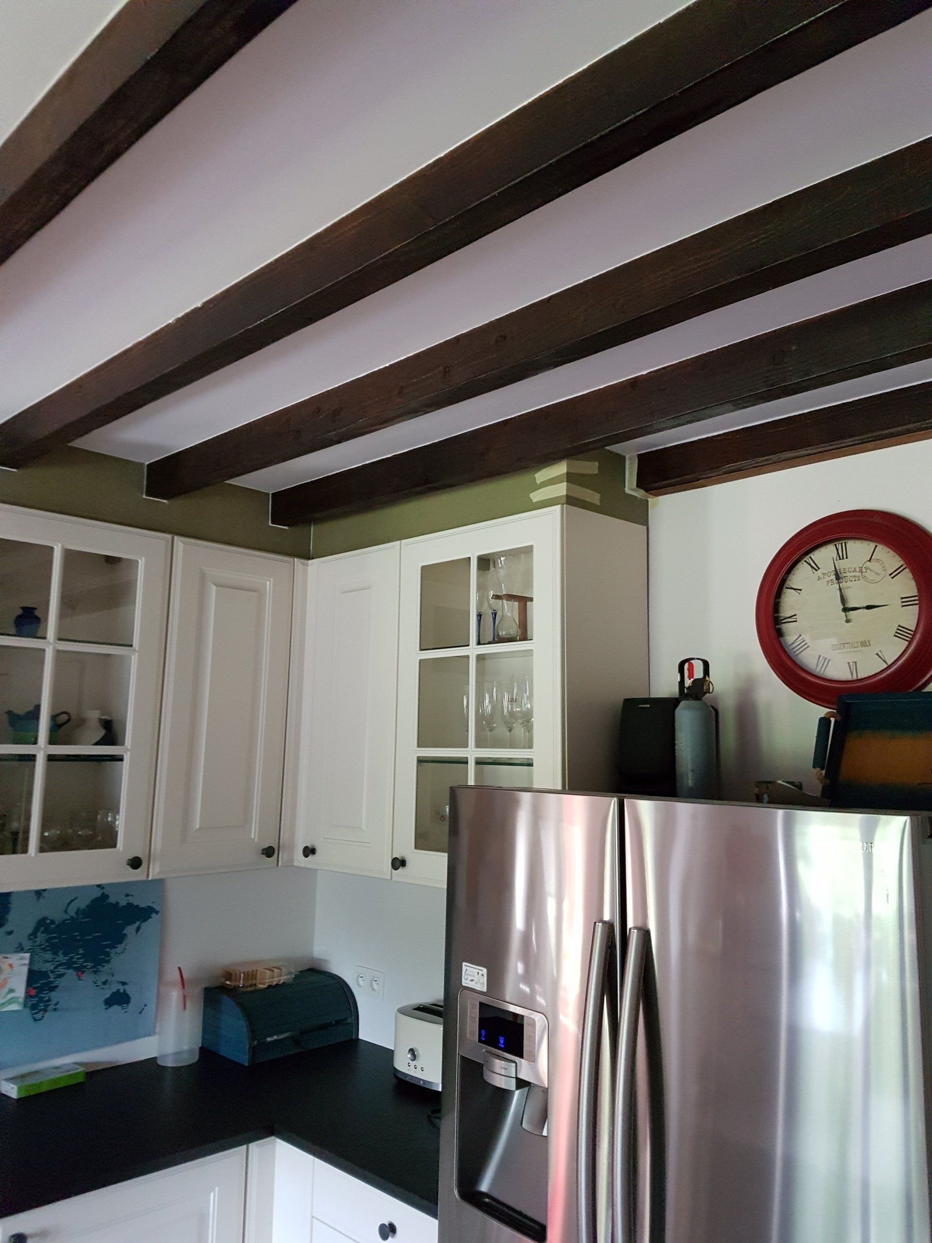 Keuken met witte kasten, een roestvrijstalen koelkast, een rode klok en donkere houten balken aan het plafond.