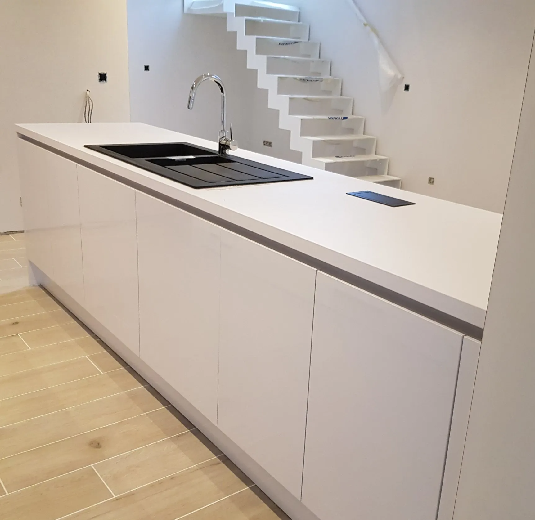 Modern wit kookeiland met zwarte spoelbak, kraan en stopcontacten, vlakbij de trap met witte stootborden en houten vloer.