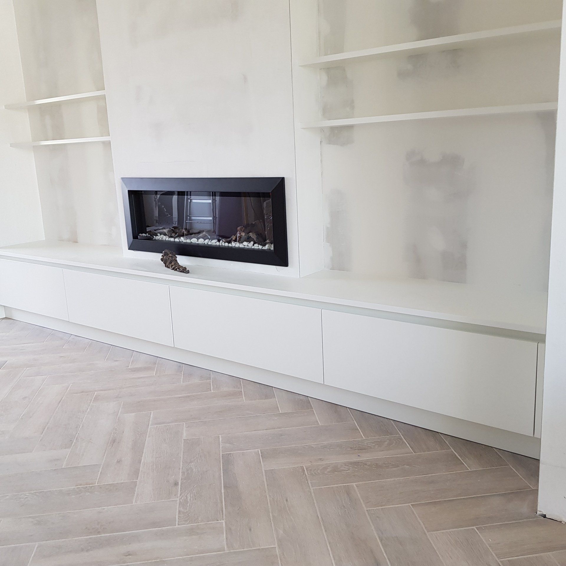 Wit ingebouwd entertainmentcentrum met open haard en planken, onafgewerkte constructie, visgraatvloer.