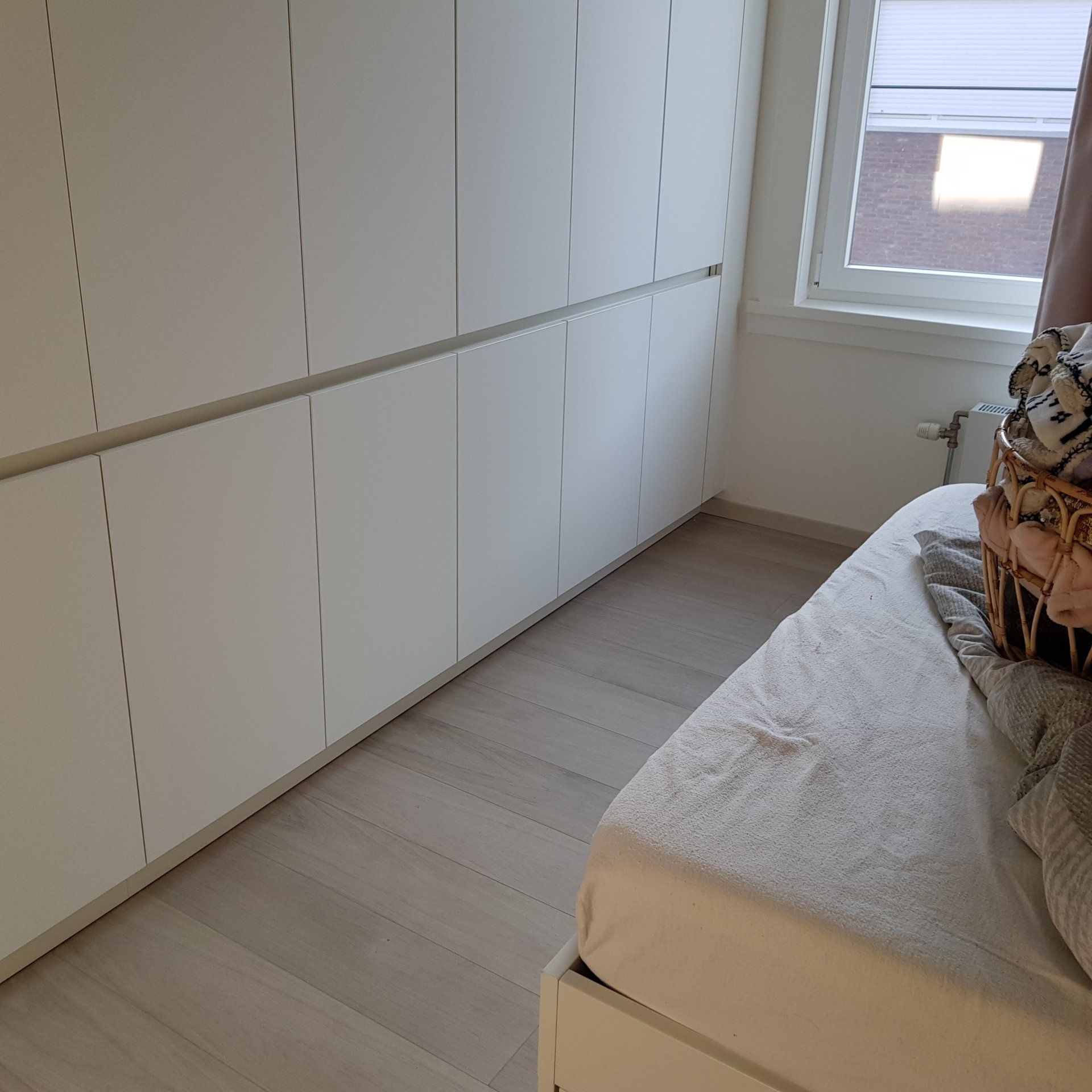 Een slaapkamer met witte kastdeuren, een bed en een raam. De vloer is van licht hout.