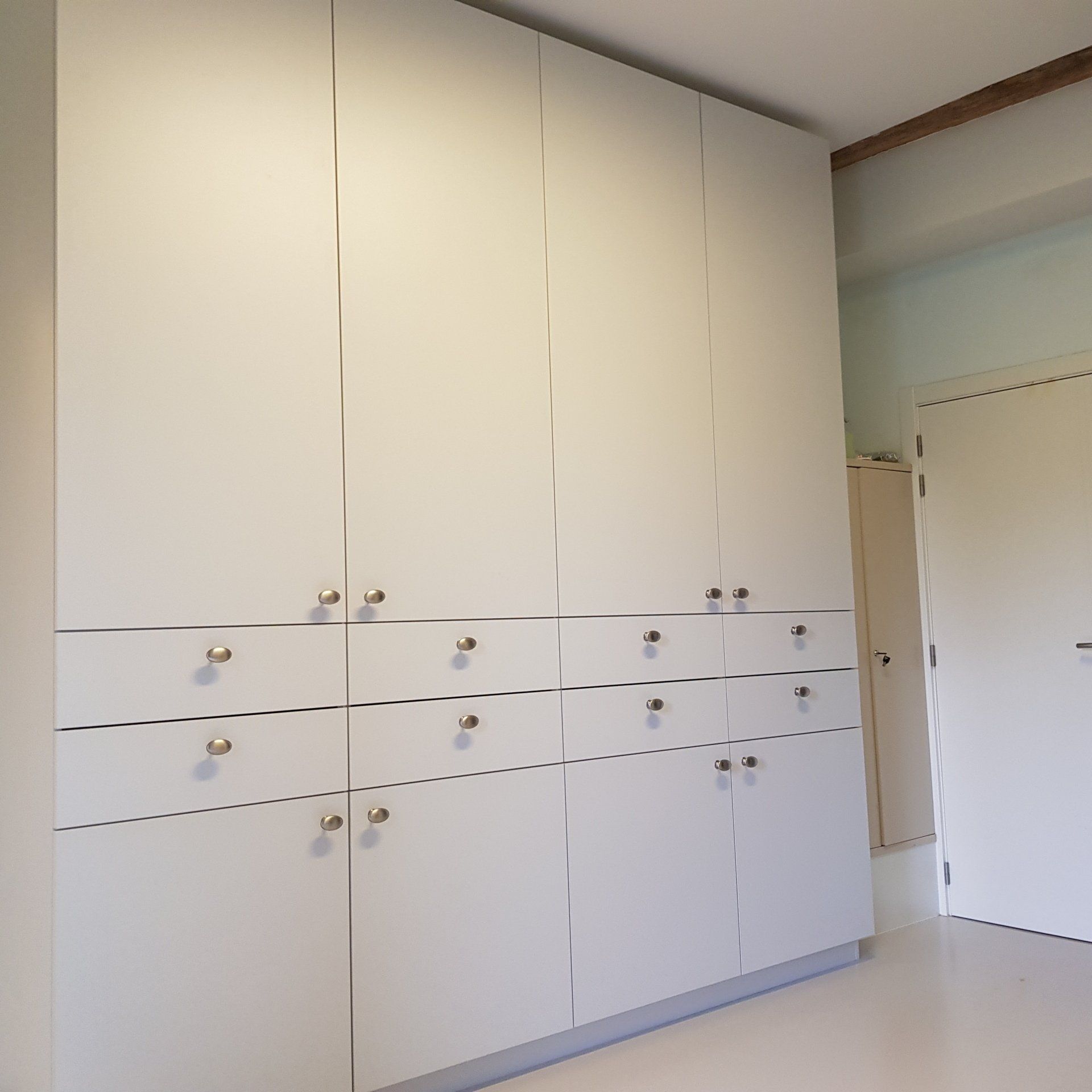 Witte inbouwkast met meerdere deuren en lades, voorzien van zilveren knoppen. De kast bevindt zich in een kamer met lichtgekleurde muren en een deur.