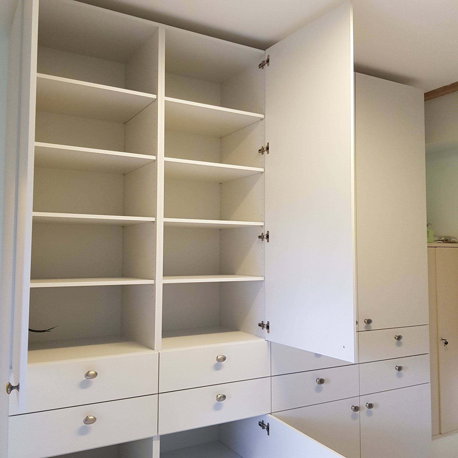 Witte inbouwkast met open planken en lades. Eén deur staat open, waardoor er lege opbergruimte zichtbaar is.