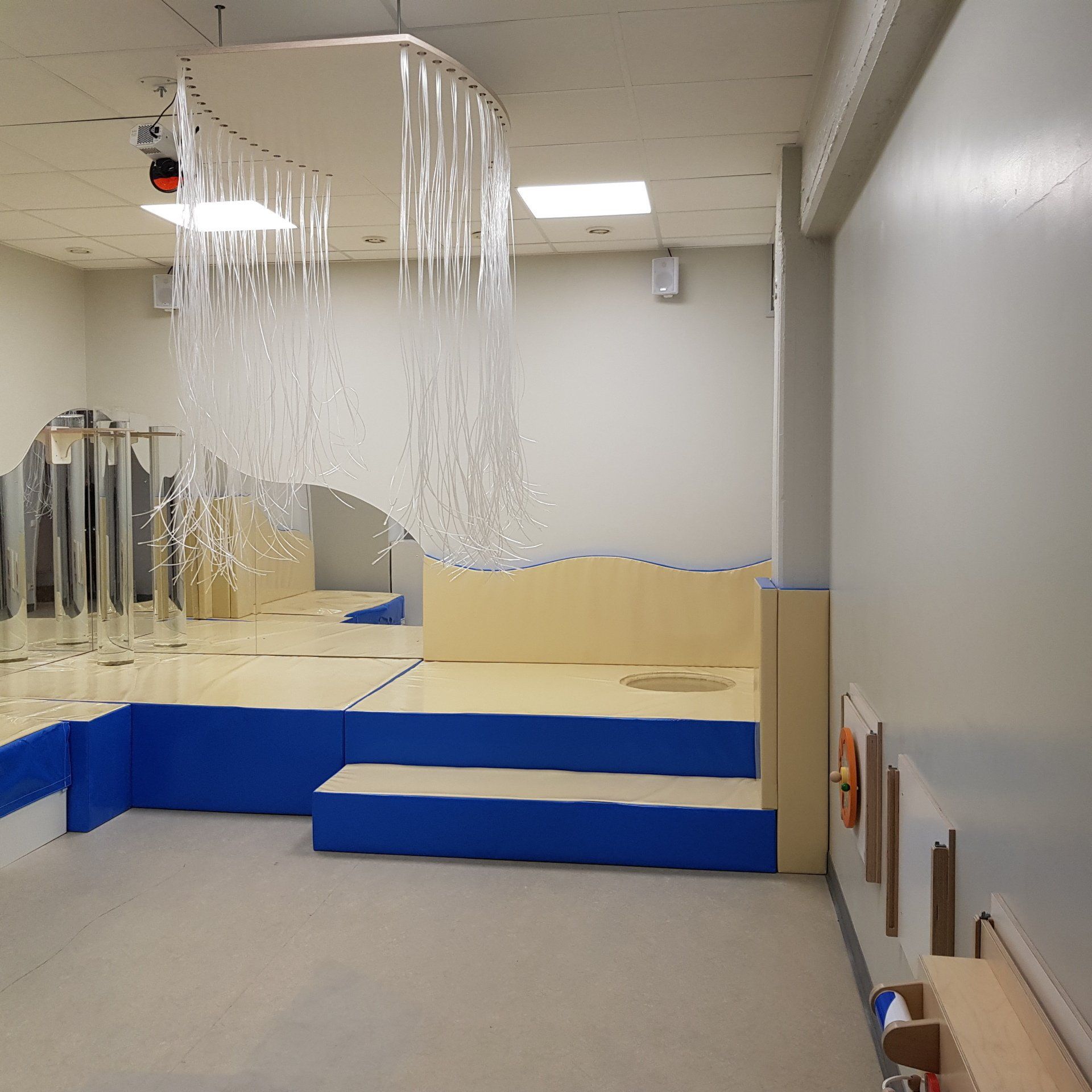 Een toegankelijke zintuigenkamer met blauwe treden, spiegels en bungelende doorzichtige linten. Een reddingsboei is aan de muur bevestigd.