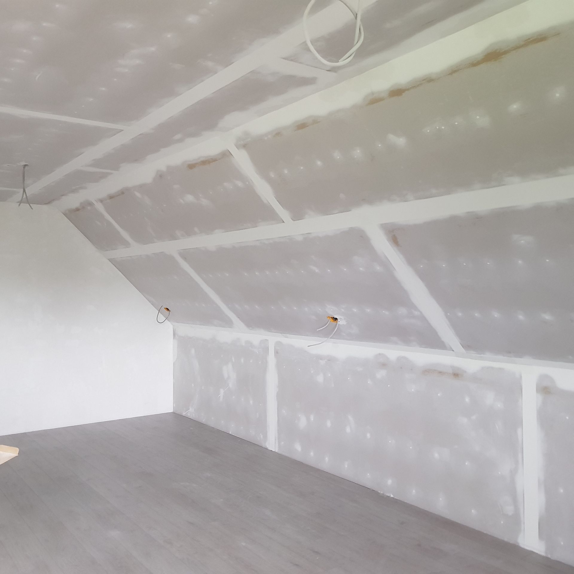Interieur van een kamer in aanbouw; gipsplaten geïnstalleerd op het schuine plafond en de muur, met grijze vloer.