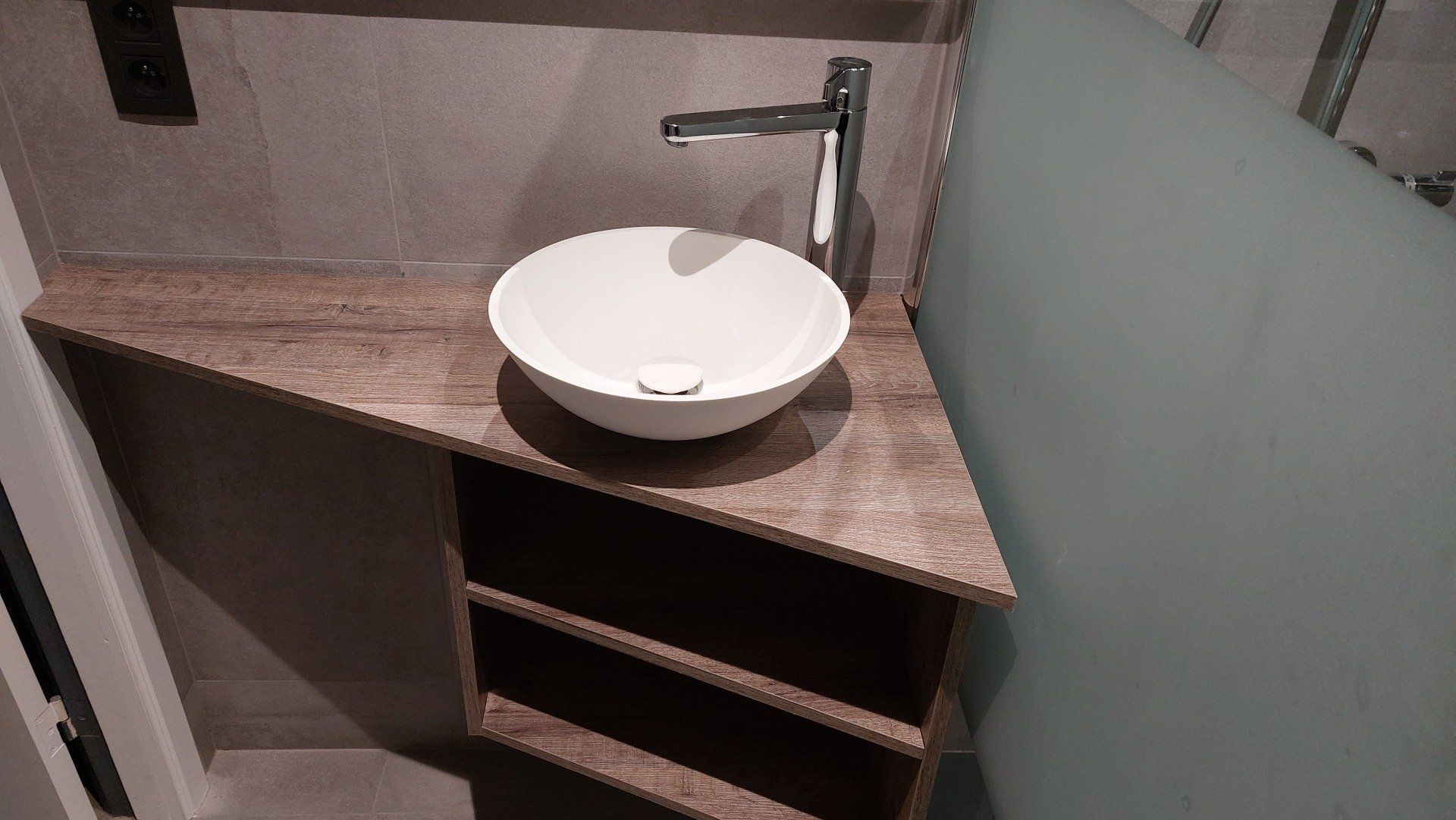 Een moderne badkamerhoek met een witte wastafel op een bruin aanrecht en een metalen kraan. Daaronder bevindt zich een opbergkast.