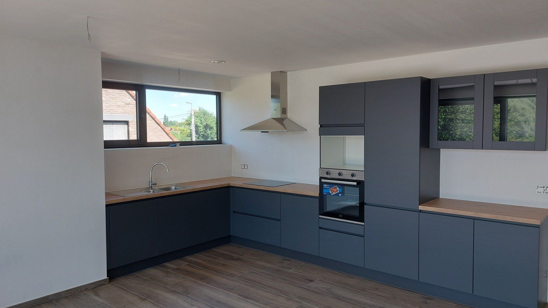 Moderne keuken met grijze kasten, houten aanrechtbladen en een roestvrijstalen afzuigkap. Een raam biedt uitzicht naar buiten.