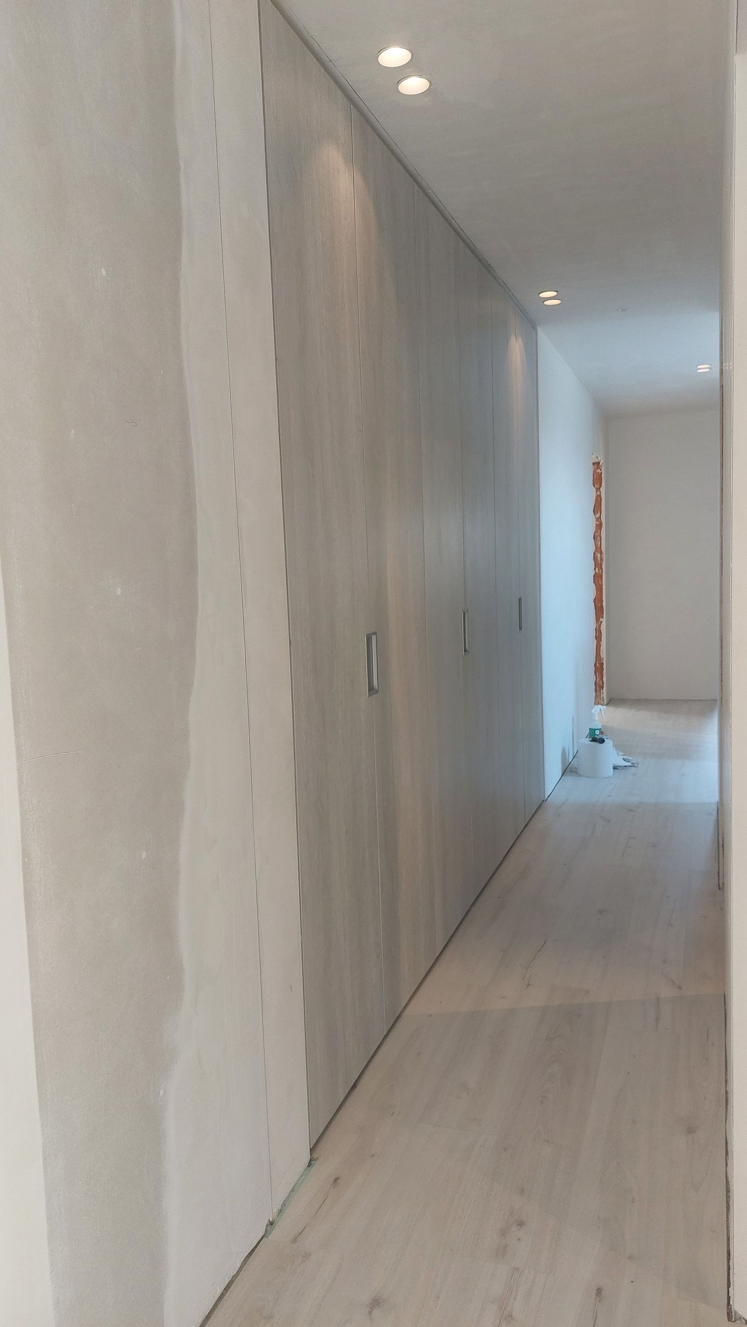 Smalle gang in aanbouw met grijze gipsplaatwanden en een lichte vloer; inbouwplafondlampen.