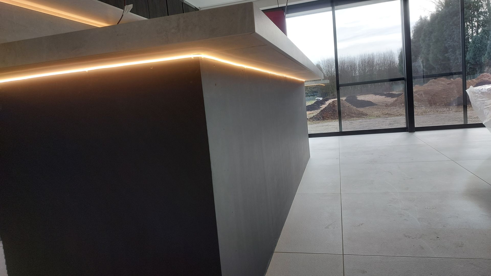 Modern kookeiland met lichtstrip, grijze en witte oppervlakken en een groot raam met uitzicht op de buitenruimte.