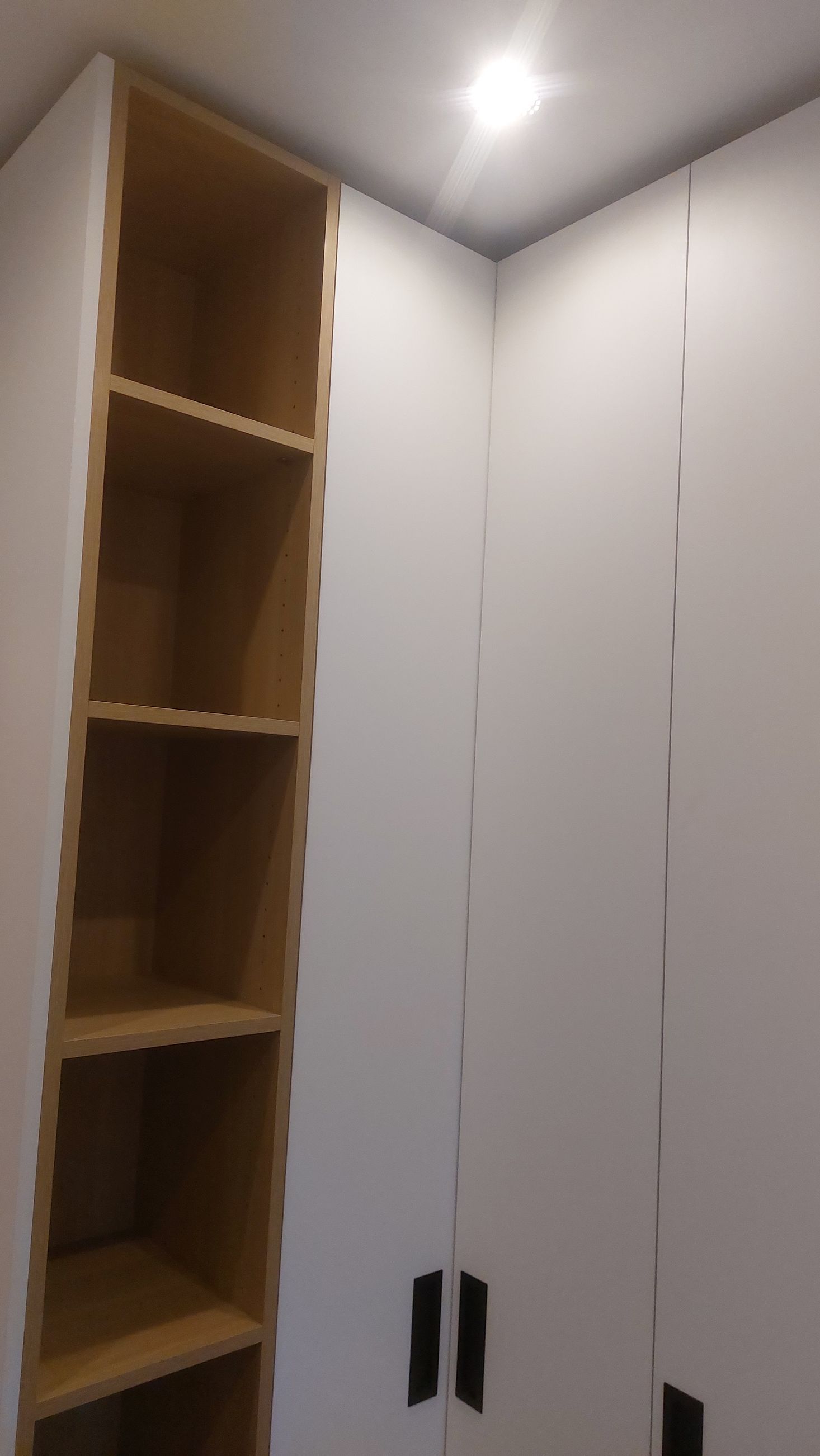 Ingebouwde hoekkast met open houten planken aan de linkerkant en witte kastdeuren aan de rechterkant, onder een inbouwplafondlamp.