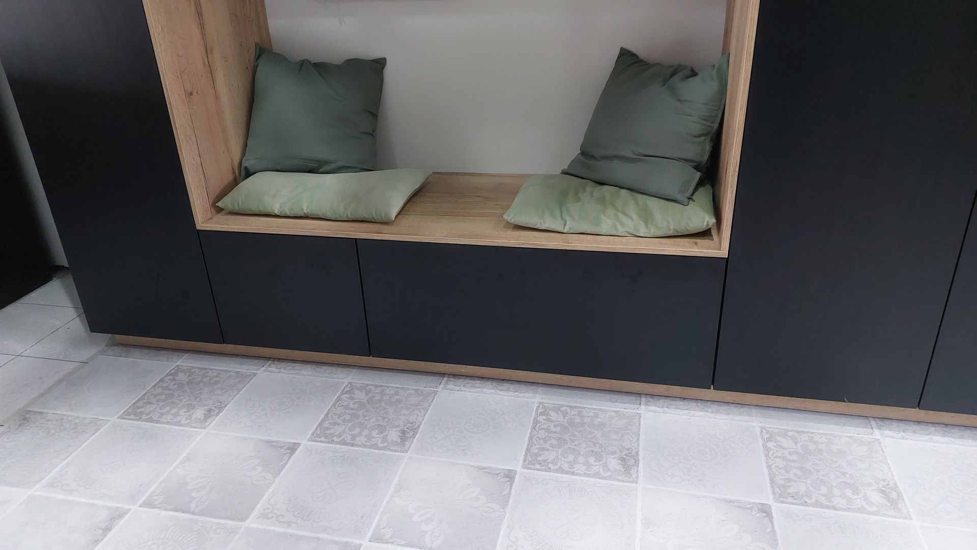 Ingebouwde zithoek met houten frame, donkere kasten en groene kussens. Grijze tegelvloer.