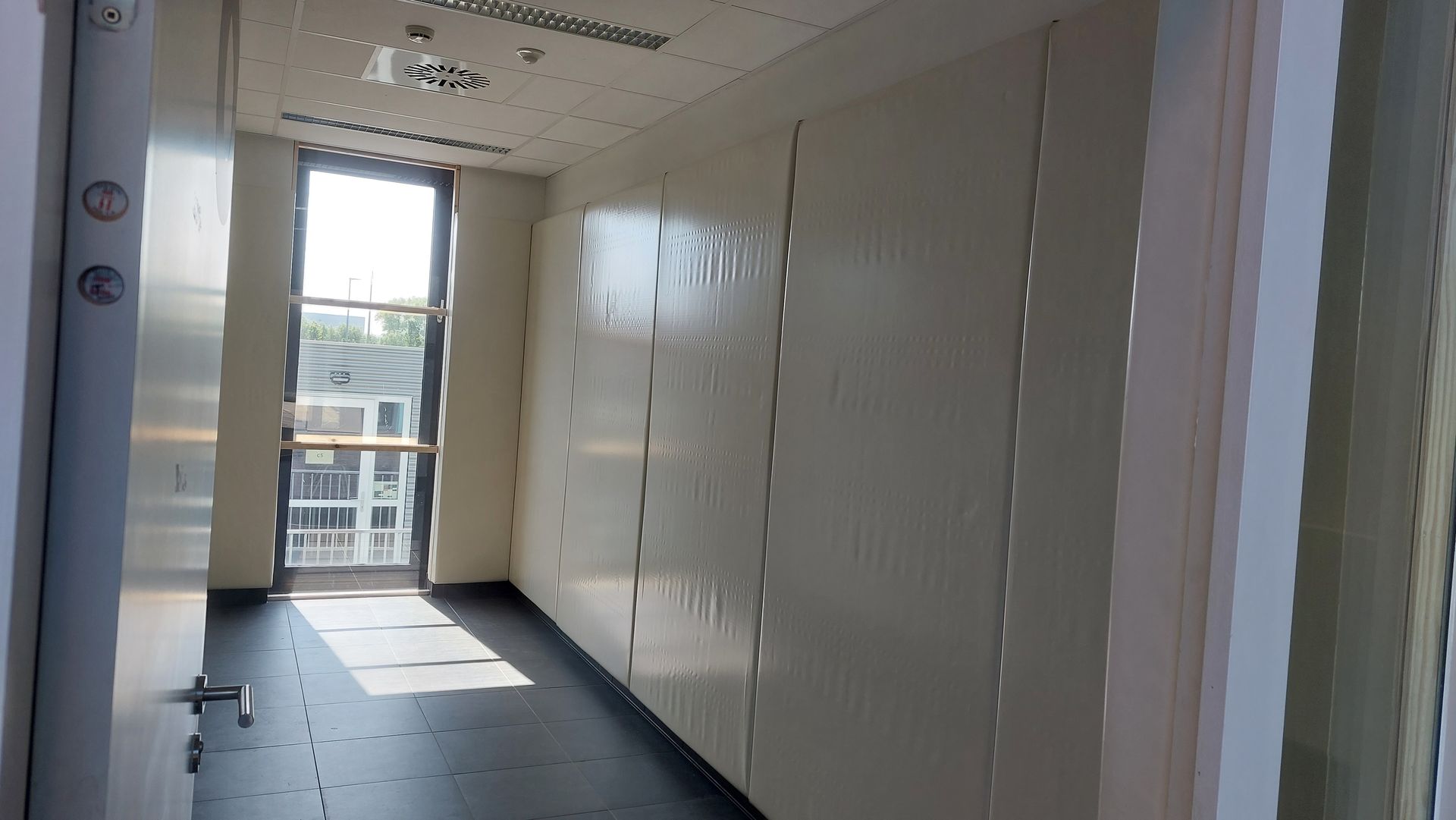 Binnenaanzicht van een smalle kantoorruimte met witte muren en een raam met uitzicht op een buitenruimte. Zonlicht stroomt de kamer binnen.