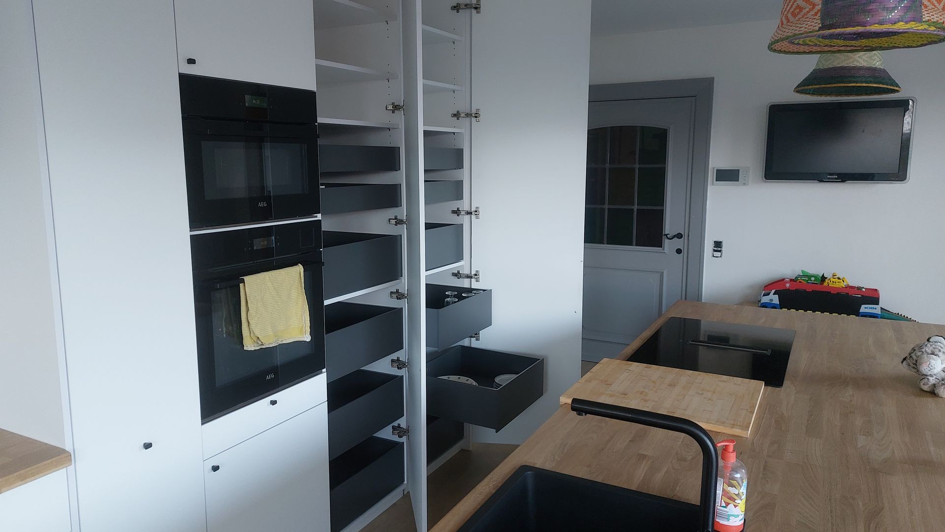 Moderne keuken met ingebouwde ovens, voorraadkast met uittrekbare lades en een houten kookeiland met spoelbak. Er hangt een tv aan de muur.