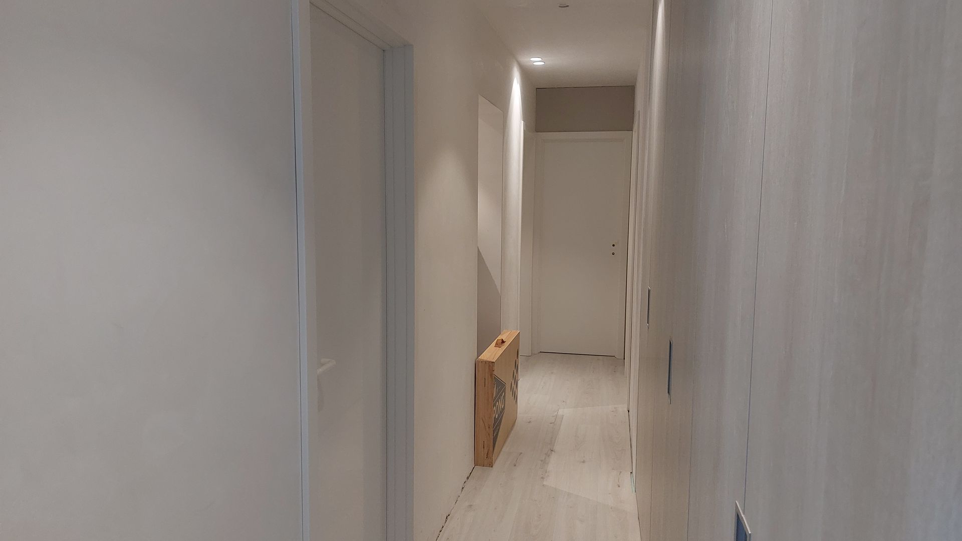 Een smalle gang met witte muren en een witte deur aan het einde. Een kartonnen doos staat tegen een muur.