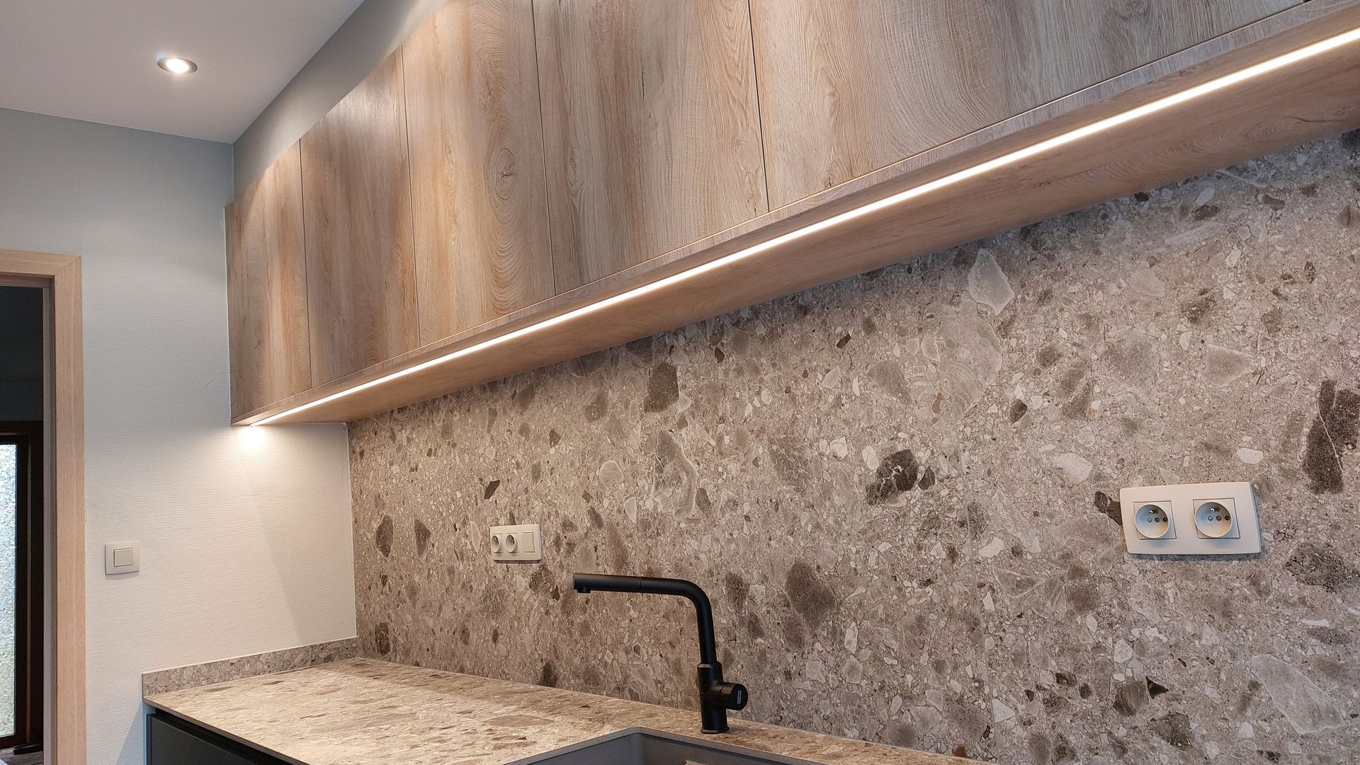 Keuken met stenen achterwand, granieten aanrechtblad en houten kasten met plafondverlichting. Zwarte kraan en stopcontacten zijn zichtbaar.