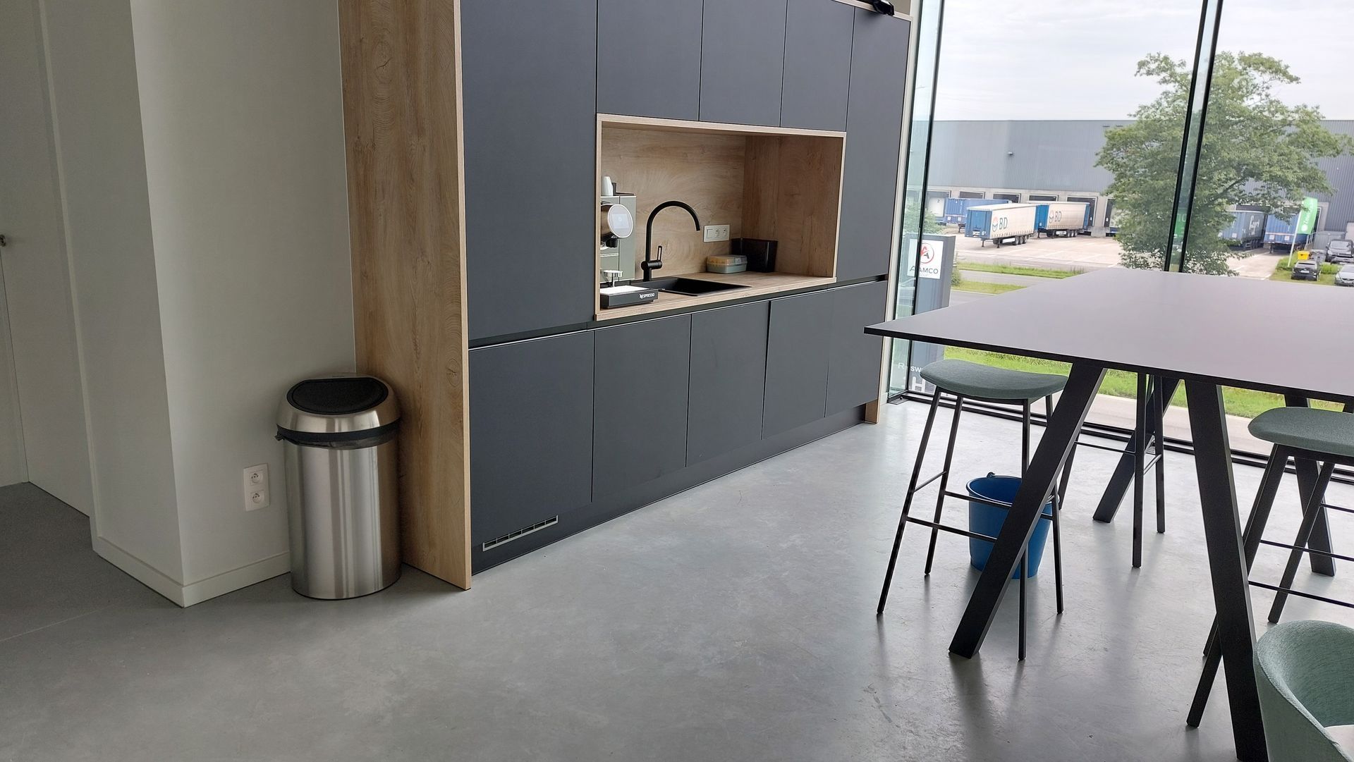 Moderne kantoorkeuken met donkere kasten, spoelbak en een tafel met stoelen. Links staat een prullenbak en een raam kijkt uit op een magazijn.
