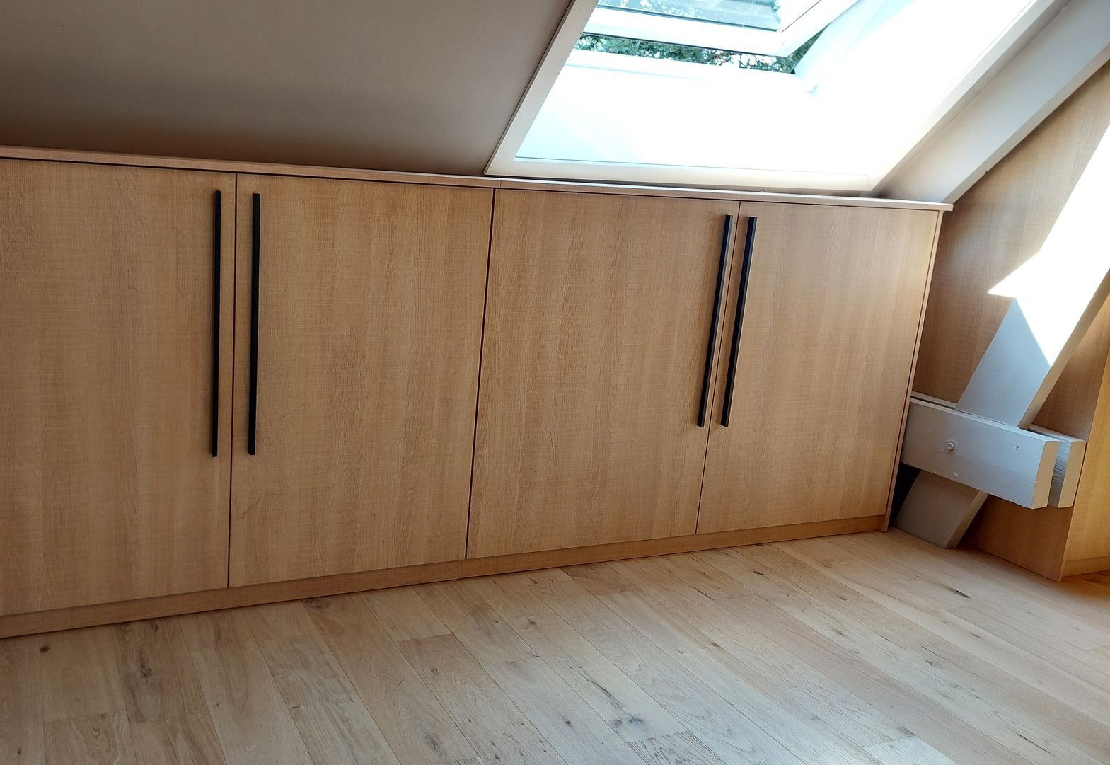 Houten opbergkasten geïnstalleerd onder een schuin plafond met een dakraam. Zwarte handgrepen op de deuren en een houten vloer.