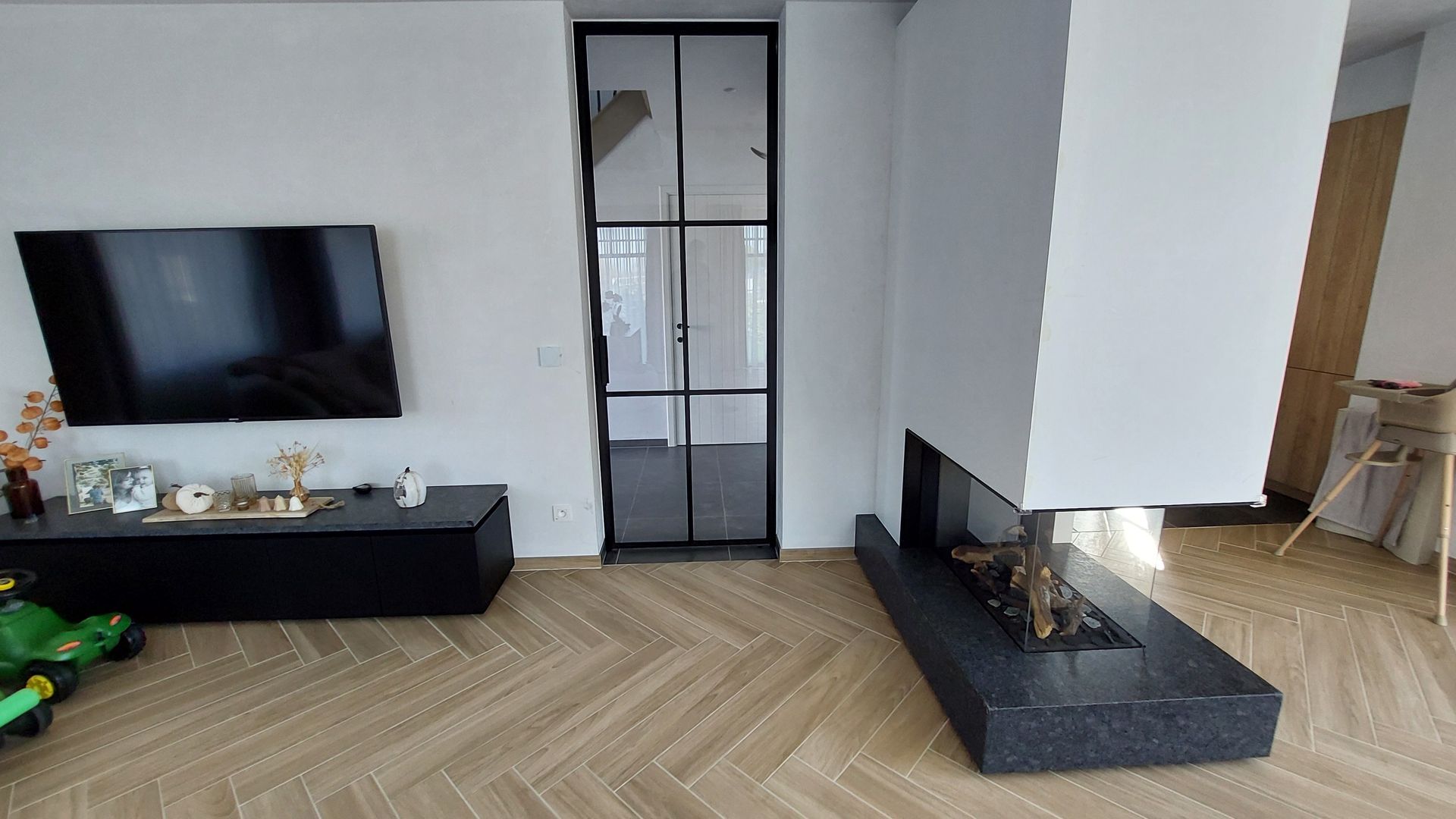 Moderne woonkamer met visgraatvloer, zwart tv-meubel, open haard en zwarte glazen deur.