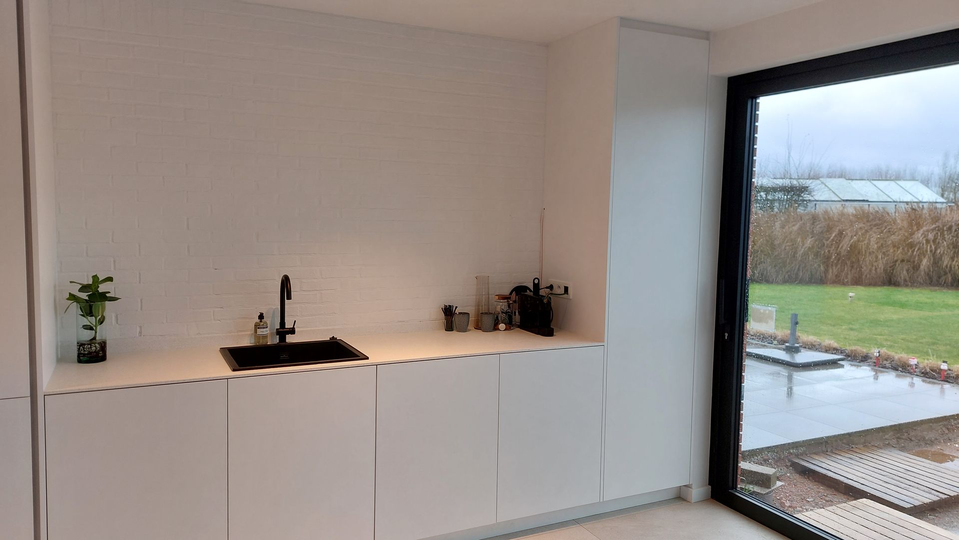 Witte keuken met een zwarte spoelbak en kraan, kastdeuren en een groot raam met uitzicht op een gazon.