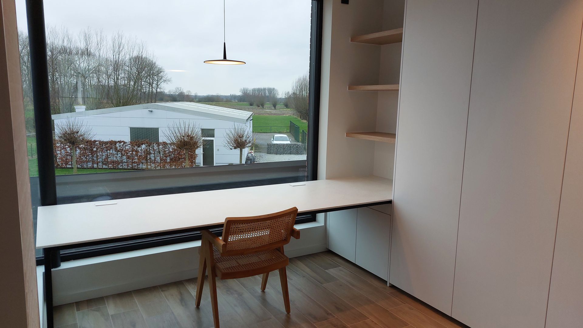 Een moderne bureauopstelling met een groot raam. Wit bureau en kasten met een lichtbruine stoel.